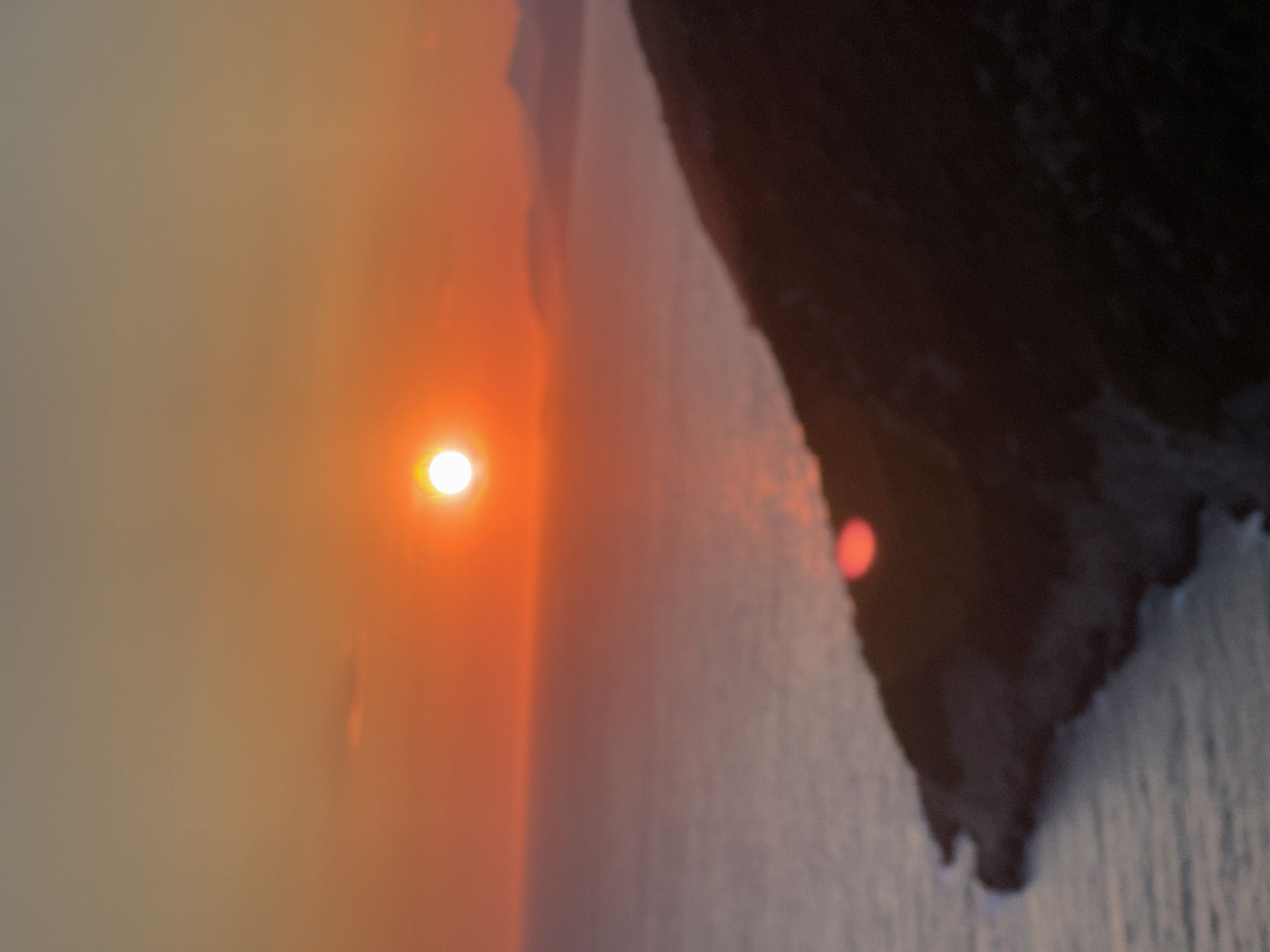 A blurred, wide-angle photograph captures a sunset over the Adriatic Sea, with a mountainous coastline of Zagora, Montenegro, in the foreground. The sun, a bright orange orb, sits low on the horizon, casting a warm glow across the sky and water. The sea is a hazy expanse of muted blues and grays, with a faint reflection of the sun visible on its surface. The coastline, a dark silhouette against the vibrant sky, rises from the sea, its rugged terrain suggested by the textured blur. Distant, hazy mountains can be seen in the background, adding depth to the scene. The overall impression is one of serene, natural beauty, with the intense colors of the sunset dominating the composition.