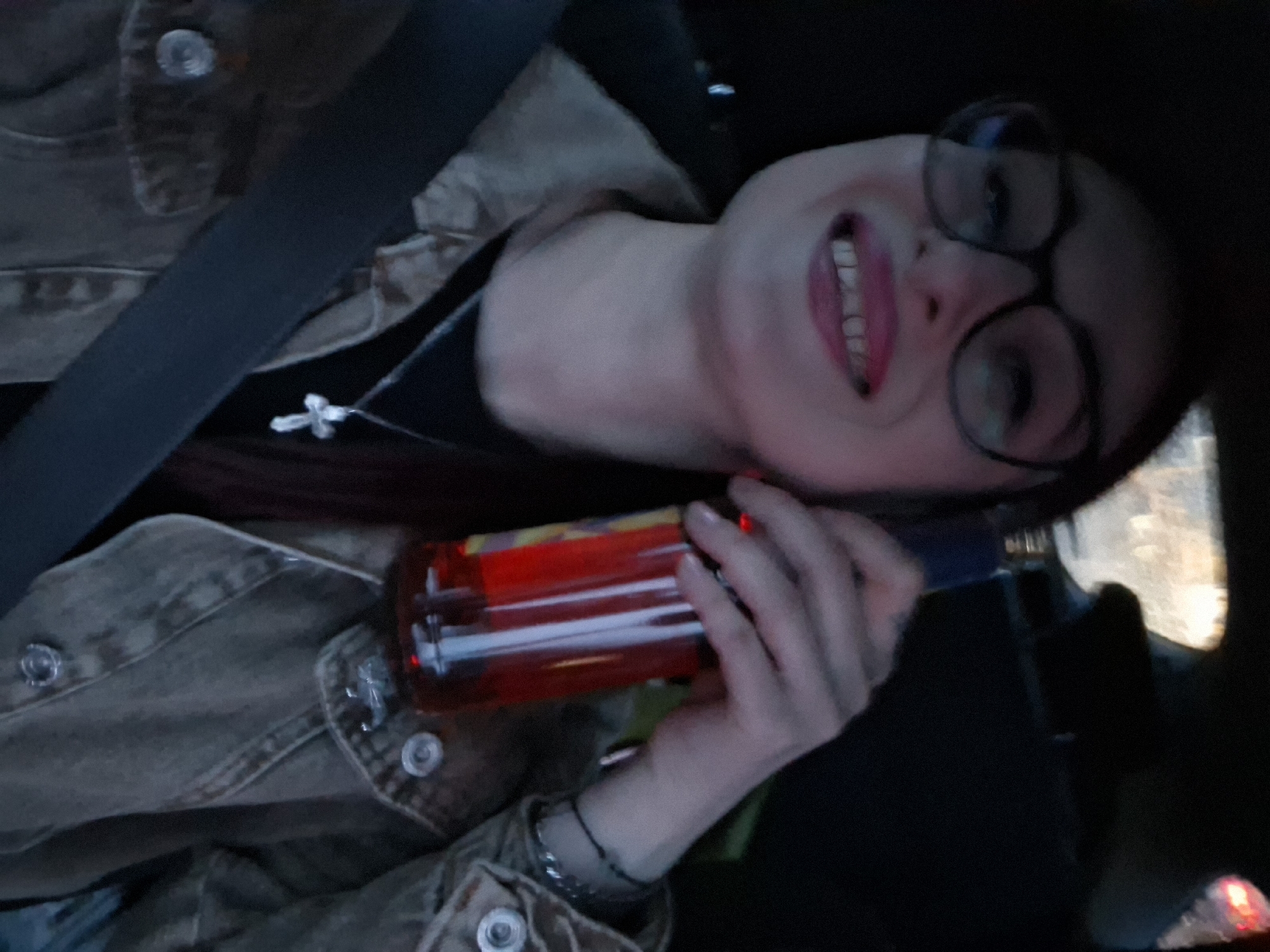 A person is shown in a low-light, indoor setting, likely a vehicle. The individual has light skin, dark hair, and is wearing glasses, a black shirt, and a buttoned jacket with a cross pendant necklace. The person is smiling with their tongue slightly out. The person is also holding a cylindrical object containing a red liquid. A seatbelt is visible over the person's left shoulder. The location is unspecified.