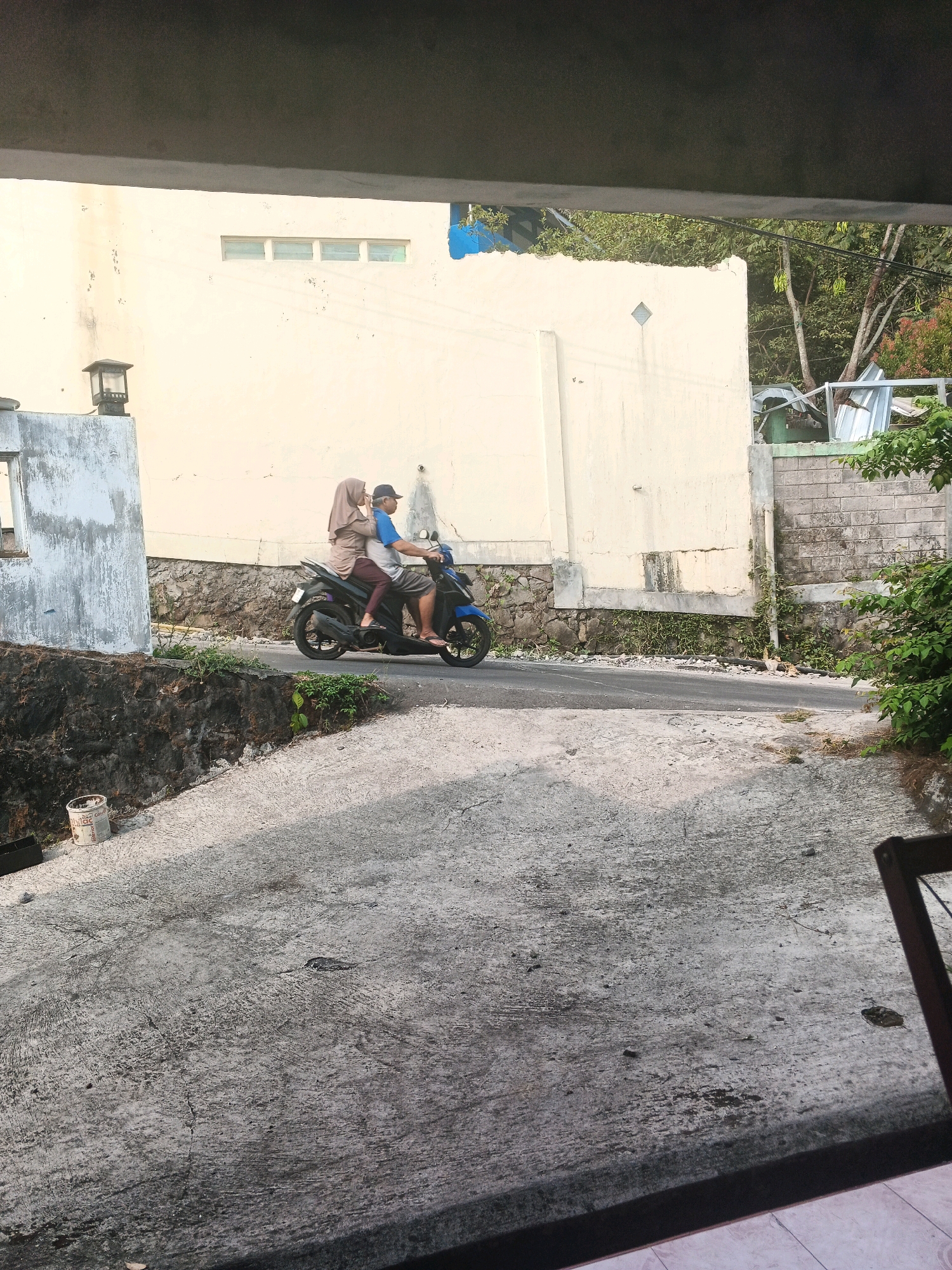 In Keji, Indonesia, a man and a woman ride a blue and black scooter on a paved road. The man, wearing a blue shirt and a dark cap, drives the scooter. The woman, in a light brown hijab and dark pants, sits behind him. The road is bordered by a concrete wall and a stone retaining wall with plants growing on it. In the background, a light-colored building with a row of windows and a blue accent is visible, along with trees and vegetation. The foreground shows a textured concrete surface, possibly a driveway or pathway, with some debris scattered on it. A white bucket with writing on it is also present in the foreground.