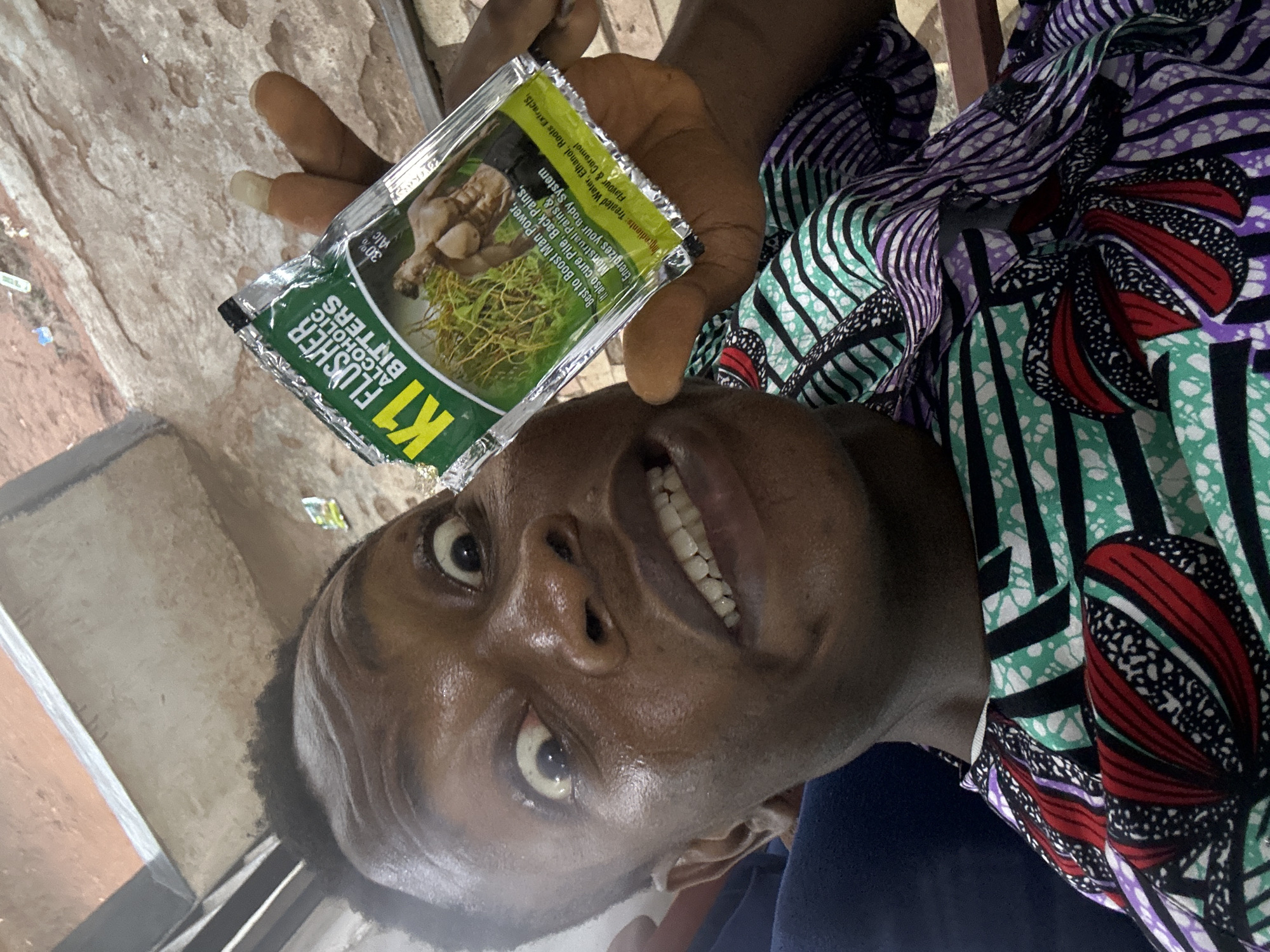 A person is holding a packet of "K1 Alcoholic Bitters." The packet claims it is "best to boost man power." The person is smiling. They are wearing a brightly patterned shirt. The location is unknown.