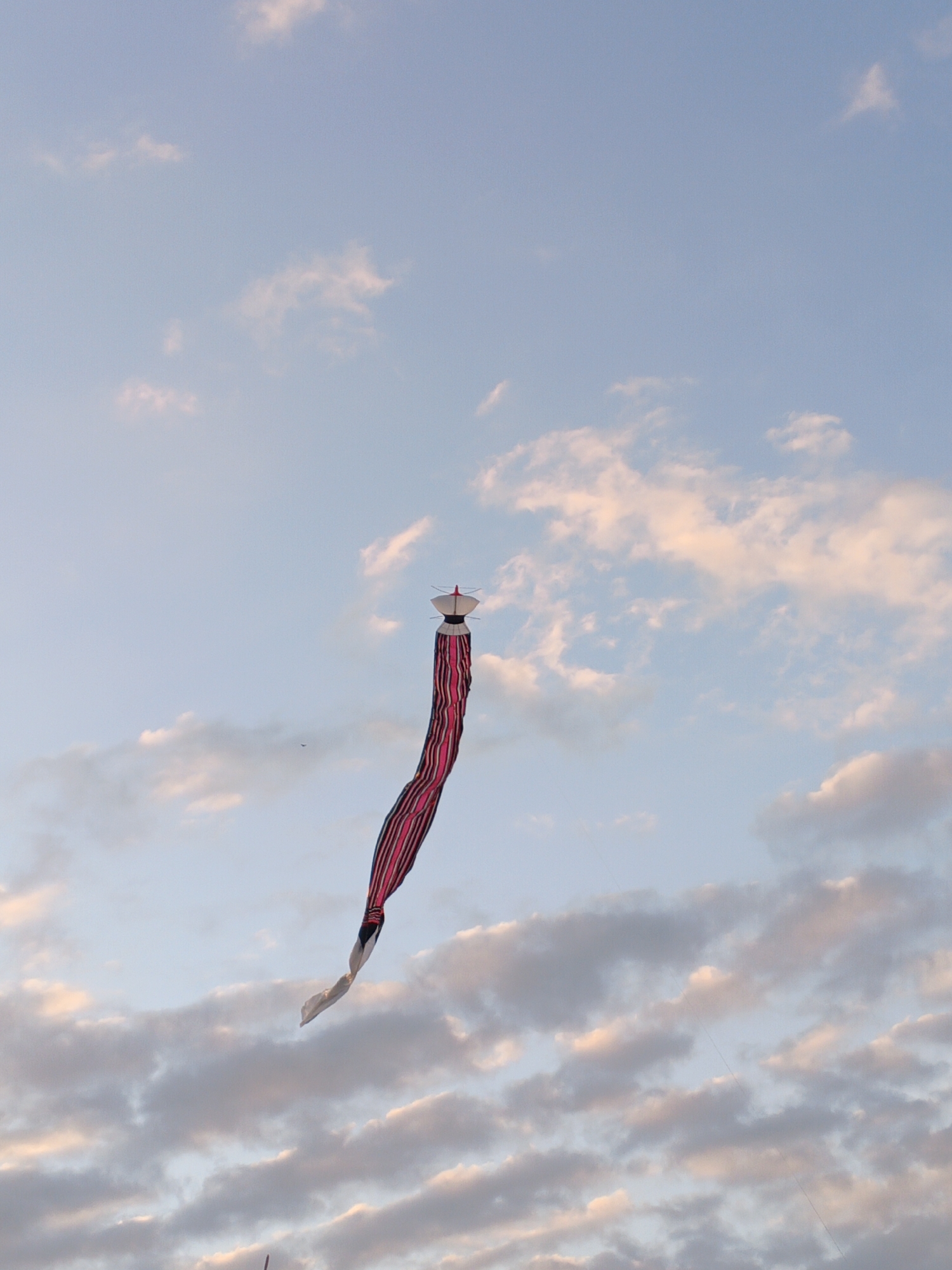 A traditional Balinese kite, known as a "layangan," is captured in flight against a pale blue sky dotted with soft, wispy clouds. The kite features a white and red diamond-shaped head with a pointed top, from which a long, flowing tail descends. The tail is made of fabric with bold, alternating stripes of vibrant pink and dark red, creating a visually striking effect as it waves in the wind. The kite is positioned in the upper right quadrant of the image, angled downwards. The background is dominated by the sky, with a scattering of clouds that are tinged with the warm hues of either sunrise or sunset. The overall scene conveys a sense of freedom and movement. This photograph was taken in Sukawati, Indonesia, a region renowned for its traditional kite-making and flying culture.