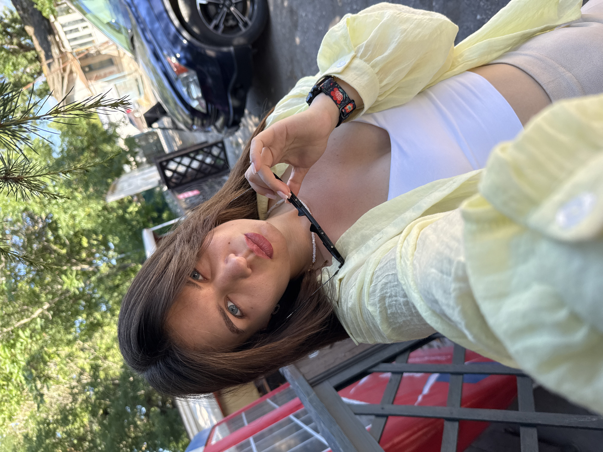 A young woman with long, dark brown hair is taking a selfie outdoors on a sunny day. She is wearing a light yellow open-buttoned shirt over a white crop top and beige pants. She is holding a pair of black sunglasses in her right hand, and her left arm is resting on her lap. She has a watch with a floral pattern on her left wrist. In the background, there is a blue car, a building with peeling paint, a red and white structure, and trees with green foliage. The woman is looking at the camera with a slight pout.