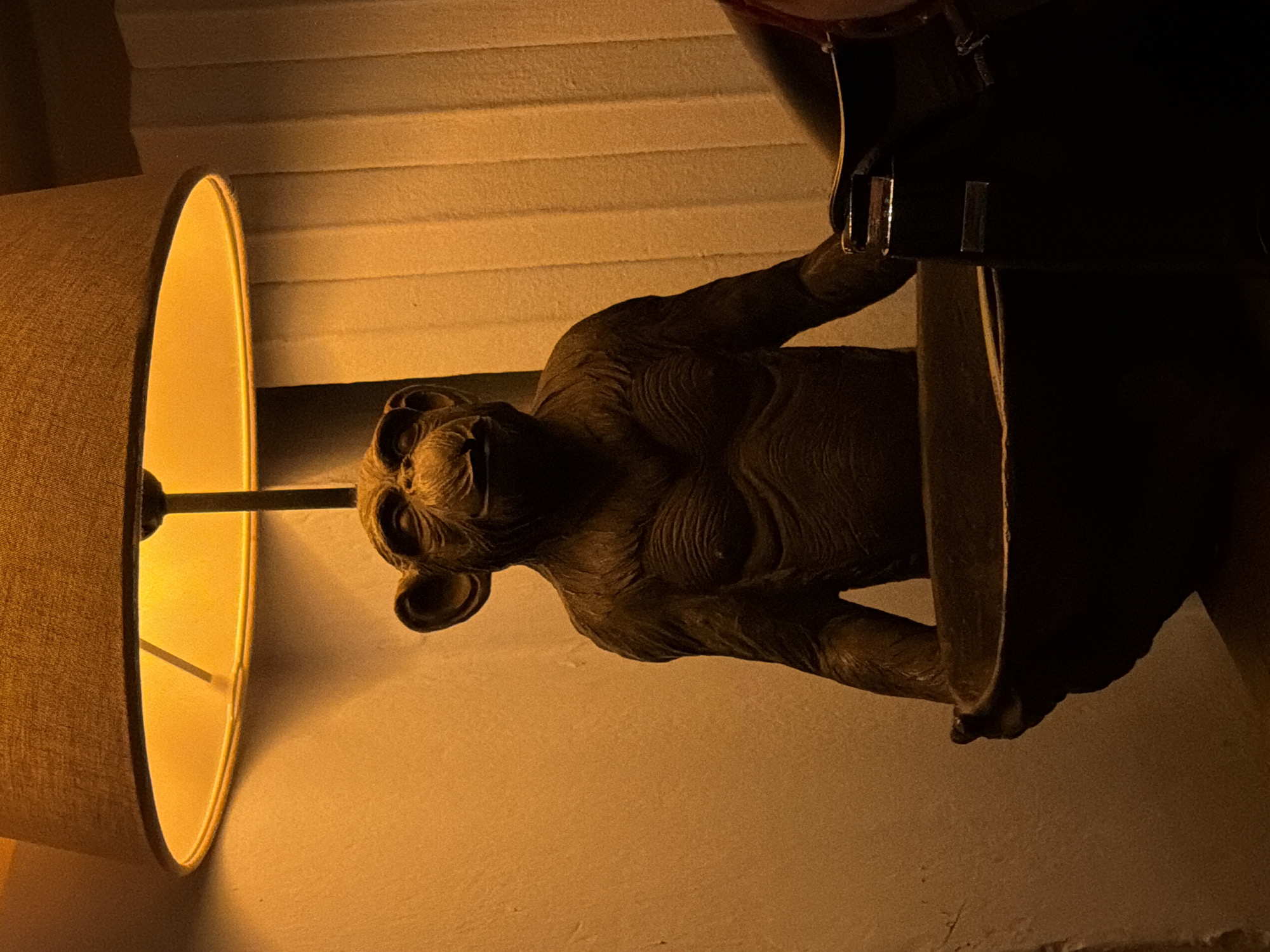 This vertically oriented image captures an atmospheric indoor scene, illuminated by warm, artificial light. On the left, a table lamp with a light brown, cylindrical fabric lampshade emits a strong, warm orange-yellow glow, casting light onto the wall behind it. The internal structure of the lampshade, including its vertical support, is visible against the bright light.
To the right of the lamp, a dark, intricately detailed statue of a creature is prominently featured. The statue appears to be made of a dark, textured material, possibly bronze or carved wood, depicting a muscular, somewhat humanoid figure with visible wrinkles and folds across its body, suggesting aged skin or bark. Its head, with prominent ears and a distinct facial structure that could be interpreted as a wide mouth or snout, is turned towards the left, partially catching the lamp's glow. The statue seems to be leaning against or holding onto an unseen object on the far right of the frame, which is mostly in shadow.
The background consists of a plain wall, bathed in the warm light from the lamp. In the upper part of the image, behind the lamp and statue, a series of light-colored horizontal slats are visible, likely part of window blinds or decorative wall paneling. The scene suggests an intimate, perhaps evening, setting, focusing on a decorative arrangement. There are no people, explicit activities, or visible text in the image. The overall impression is one of mystery and intrigue due to the unique statue and the dramatic lighting.