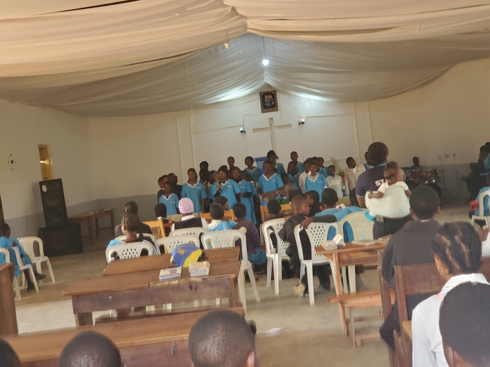 The image depicts an indoor gathering at Kuru Karama, Nigeria, likely a religious or school event. The scene is set within a room with a white, draped ceiling, creating a tent-like atmosphere. The walls are also white, and a wooden cross is mounted on the back wall, above a framed crest or emblem. Lighting appears to be artificial, with bulbs visible on the ceiling.
The main subjects are a large group of people, predominantly children and young adults, many of whom are wearing light blue shirts, suggesting a uniform or choir attire. Some adults are also present, including one woman holding a baby. The individuals are seated on white plastic chairs and wooden benches, facing towards the front of the room where a group appears to be gathered, possibly for a performance or presentation. The presence of musical instruments, including a drum set, in the background further suggests a performance or worship service. Wooden desks or tables are arranged in the foreground.
The event seems to be active and engaging, with many of the younger attendees looking towards the front. The overall atmosphere appears to be one of communal gathering and participation. There is no indication of the time of day or weather conditions. The location cues are limited to the indoor setting and the mention of Kuru Karama, Nigeria. No visible text is decipherable in the image.