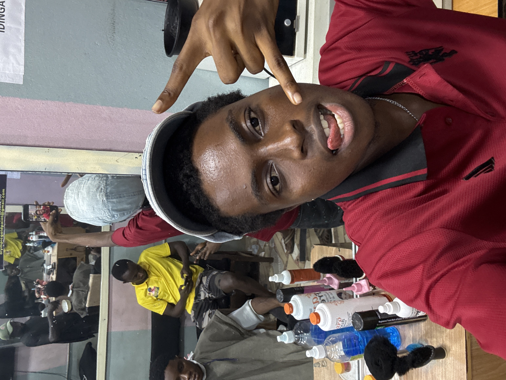The image captures a young man in a red shirt and grey cap, posing with a hand gesture. He appears to be inside a barbershop in Nkpor, Nigeria. Reflected in a mirror are several individuals, including one in a yellow shirt. Hairdressing supplies, such as bottles and hair extensions, are visible on a counter near the main subject. A sign with the email olympicgoldpaint@gmail.com is present in the background.