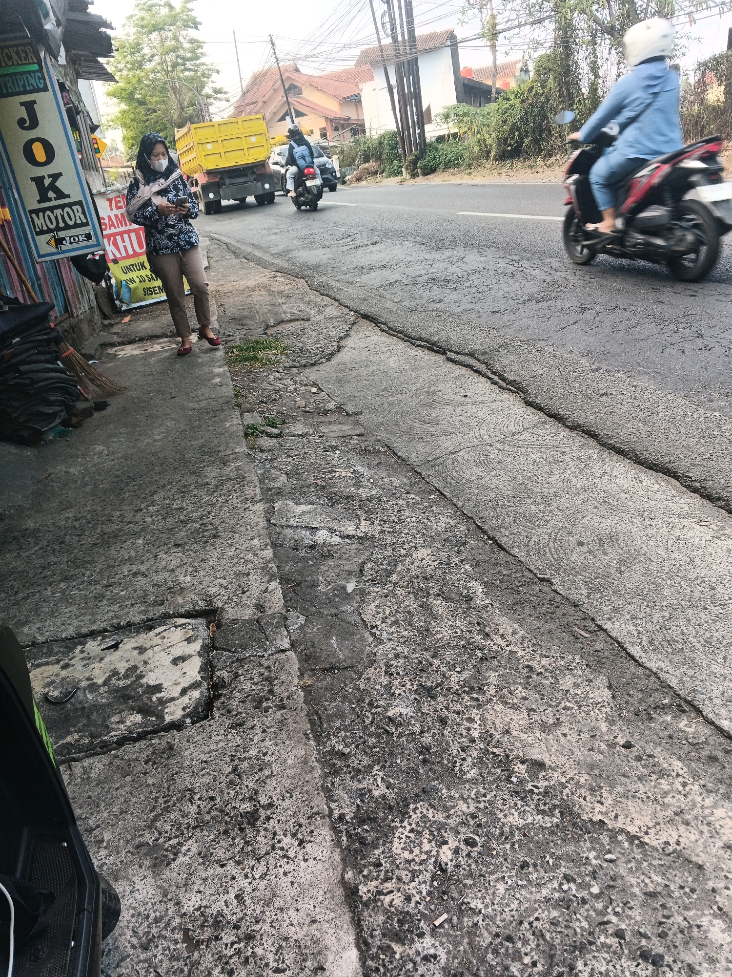 On a cracked asphalt road in Ungaran, Indonesia, a woman wearing a blue headscarf, a patterned long-sleeved shirt, and beige pants stands on a sidewalk, looking at her phone. To her left, a yellow dump truck is parked, and a broom leans against a stacked pile of black items. Across the street, a person wearing a white helmet and a blue jacket rides a scooter, moving away from the camera. The environment appears to be a roadside commercial area, with buildings and trees visible in the background.