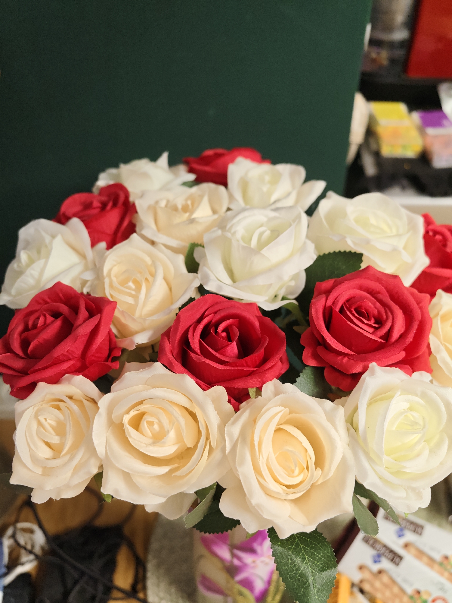 A bouquet of artificial roses, primarily in shades of red and cream, is arranged in a vase. The background is a dark green surface on the left, and a blurred, out-of-focus area on the right that suggests an indoor environment with shelves and various objects. The lighting appears to be artificial and somewhat dim. The image does not depict any recognizable landmarks or elements that would indicate a specific location within Petah Tikva, Israel.