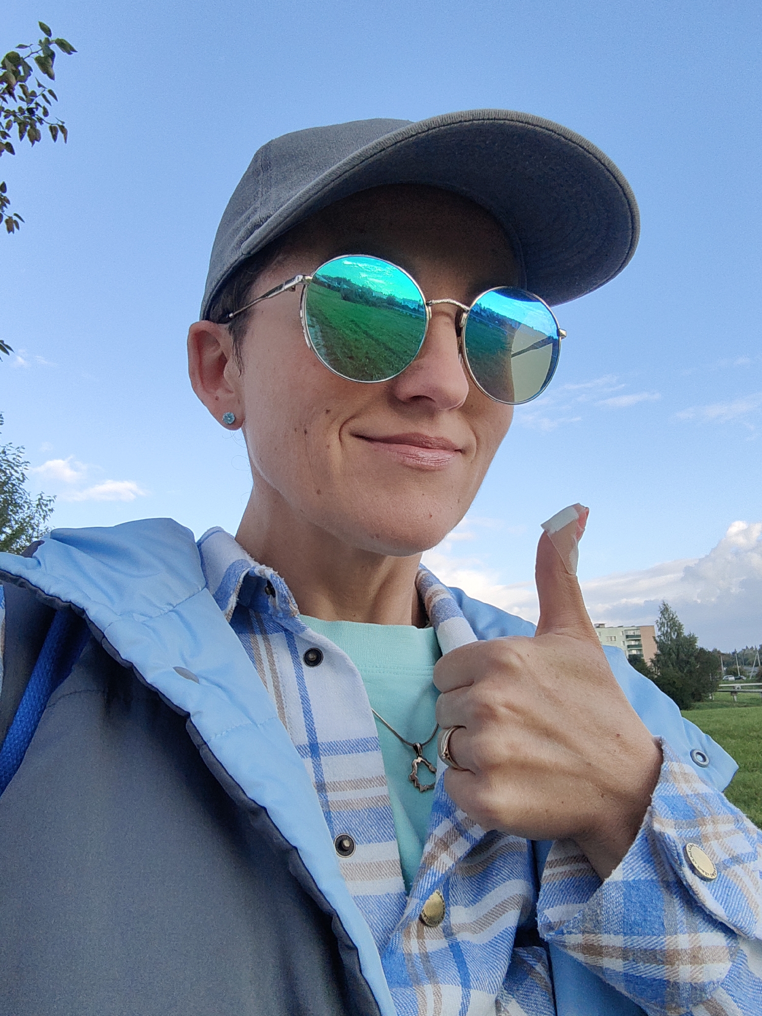 A woman with short brown hair, wearing a grey baseball cap, round mirrored sunglasses, and a light blue plaid shirt over a turquoise t-shirt, gives a thumbs-up gesture. Her right thumb is bandaged. She is outdoors with a clear blue sky and some clouds in the background, with trees and buildings visible in the distance. The scene is a park in the city of Myadzyel, Belarus.