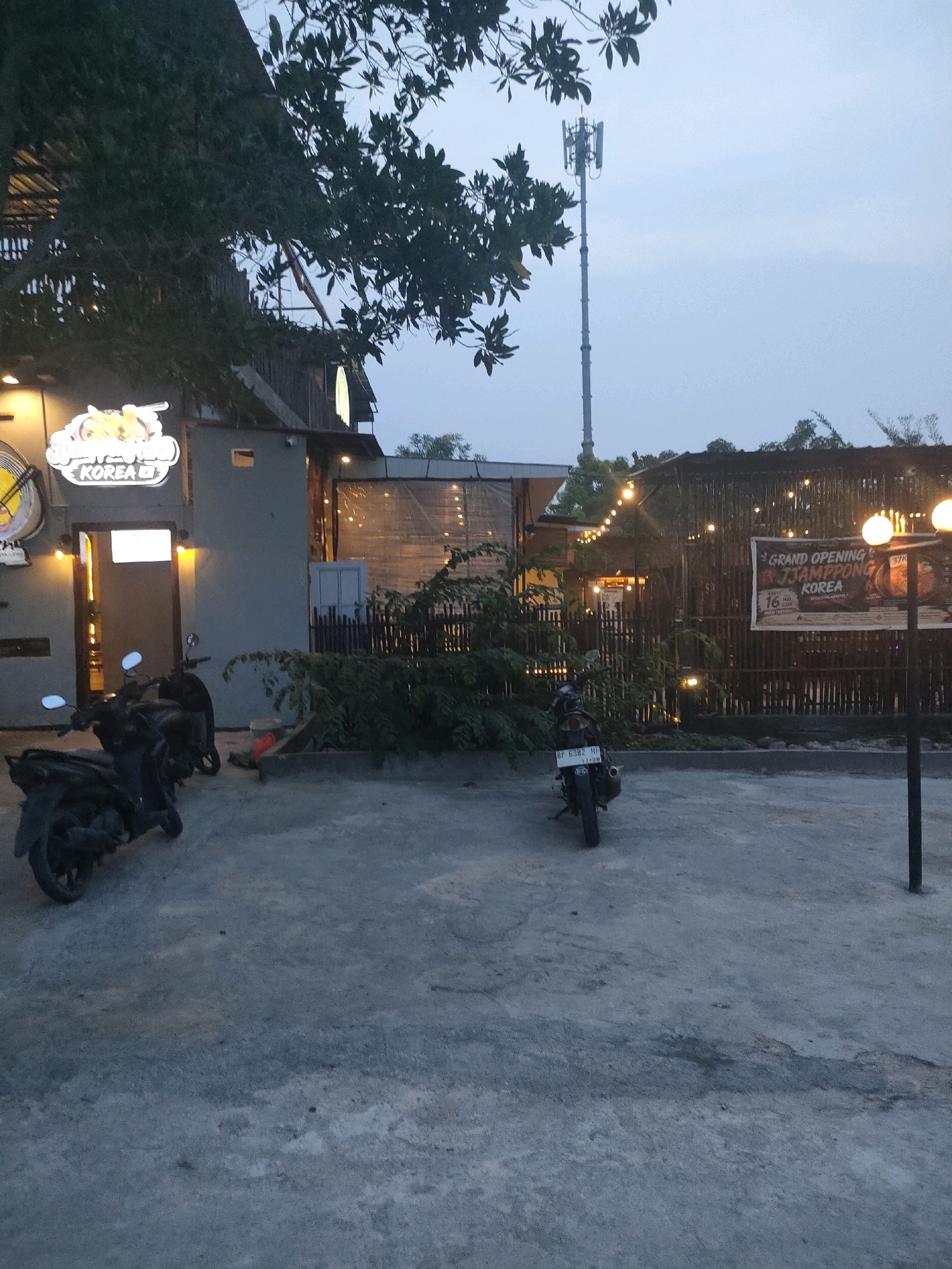 This image shows the exterior of a restaurant in Batam, Indonesia, with a prominent "Grand Opening Jjampong Korea" banner displayed on a bamboo fence. Two motorcycles are parked on a concrete surface in front of the restaurant, which has a rustic design with wooden elements and string lights. A cellular tower is visible in the background, against a dusky sky. The restaurant features a neon sign above the entrance that reads "KOREA". The overall scene suggests an evening setting, possibly just before or after sunset, with artificial lighting illuminating the area.