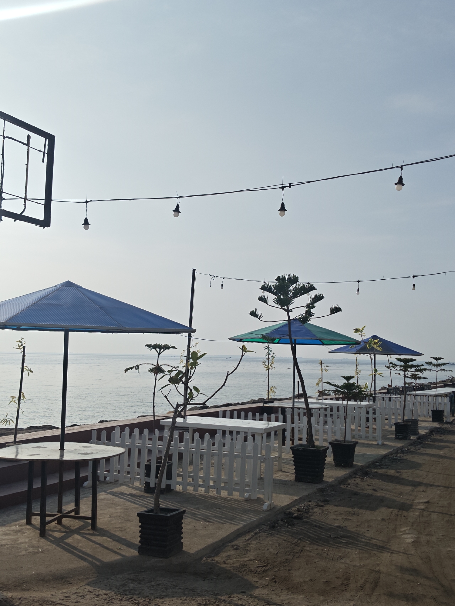 The image depicts an outdoor restaurant or seating area along a coastline. It is most likely located near the city of Lhokseumawe, Indonesia. The scene is captured during the day, under a bright, slightly overcast sky.
Several tables with white picket fences are arranged along a walkway parallel to the water. Each table appears to have an umbrella for shade; some are blue, while others are a mix of blue and green. Potted plants are placed intermittently between the tables.
A black frame-like structure and string lights are visible above the seating area, contributing to the ambiance. In the background, the ocean stretches to the horizon, with a few boats or ships visible. The ground in the foreground is a mix of concrete and dirt or sand. The overall impression is a relaxed, seaside dining area.