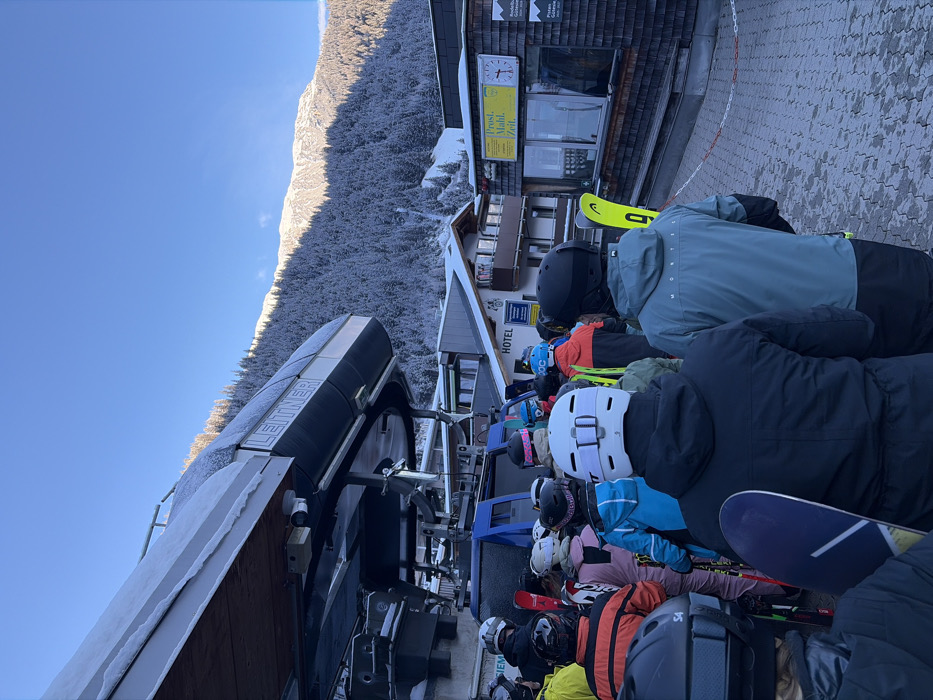 The image depicts a winter scene at a ski resort in Sankt Jakob am Arlberg, Austria, featuring a queue of approximately 15-20 individuals waiting to board a ski lift. The ski lift loading structure, branded "LEITNER" and partially covered in snow, occupies the left side of the frame. Most individuals in the queue are viewed from behind, wearing ski helmets and various colors of ski jackets and pants, including black, white, teal, orange, and blue. Several individuals are equipped with skis, some bright yellow, while at least one person in the foreground holds a purple snowboard.
The queue stands on a paved surface composed of interlocking pavers. To the right of the queue, a small, rustic building with a wooden shingle exterior is visible, featuring large windows and a sign overhead that reads "Prost. Mahl. Zeit." A clock with Roman numerals is integrated into this sign, and other partial signs below mention "Rodelbahn Galzig" and "Pisten Gelten." Behind the queue and the smaller building, a multi-story building with balconies and traditional alpine architecture is visible, bearing the word "HOTEL." The background features a large mountain covered densely with snow-laden coniferous trees, extending to snow-covered peaks under a clear, bright blue sky. Sunlight illuminates parts of the mountain, creating distinct shadow lines.