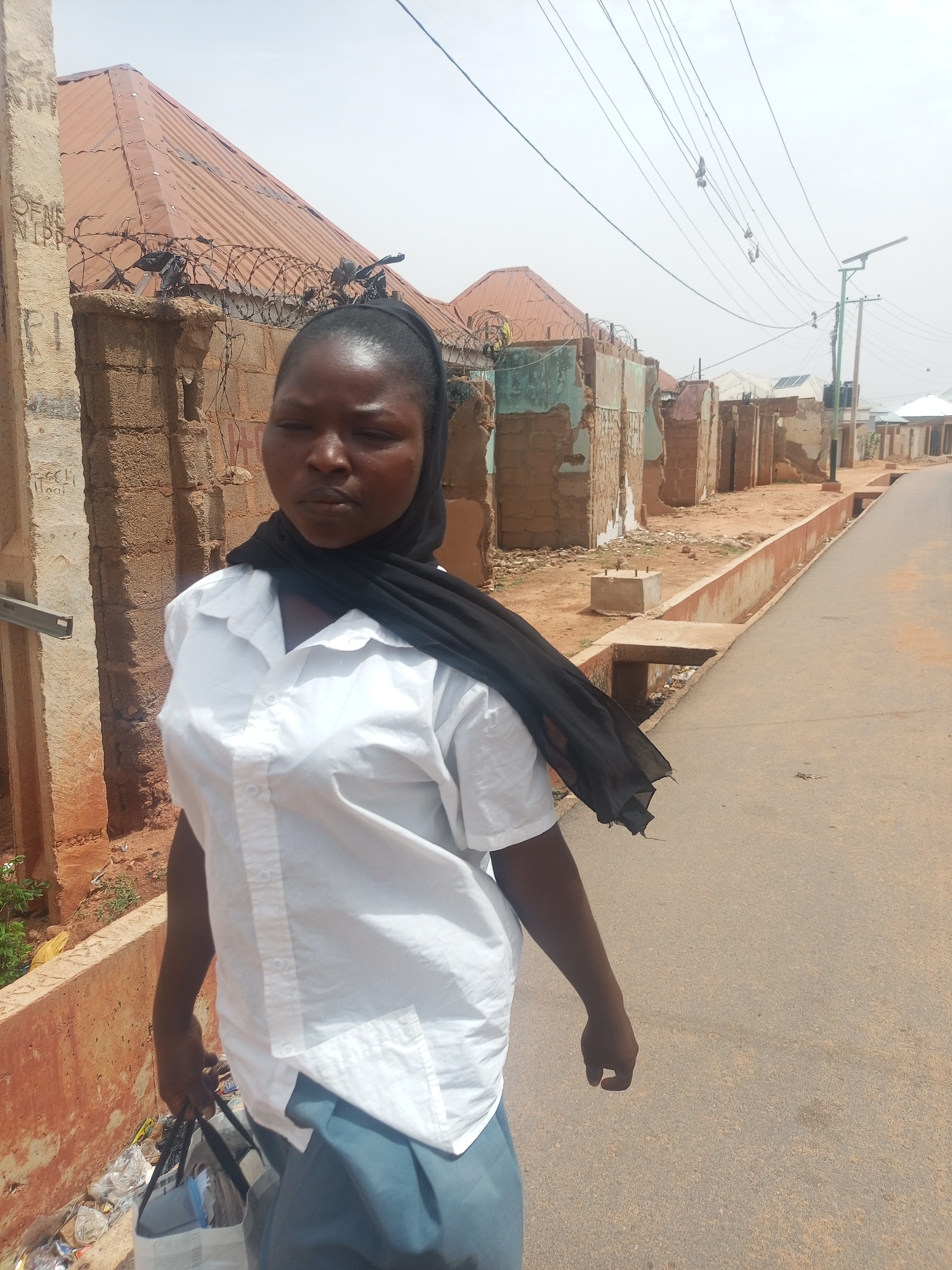 A person stands on a road in Tempure, Nigeria. The road is lined with dilapidated buildings and utility poles. The person is wearing a white shirt, a dark head covering, and carrying a bag. The sky is overcast.