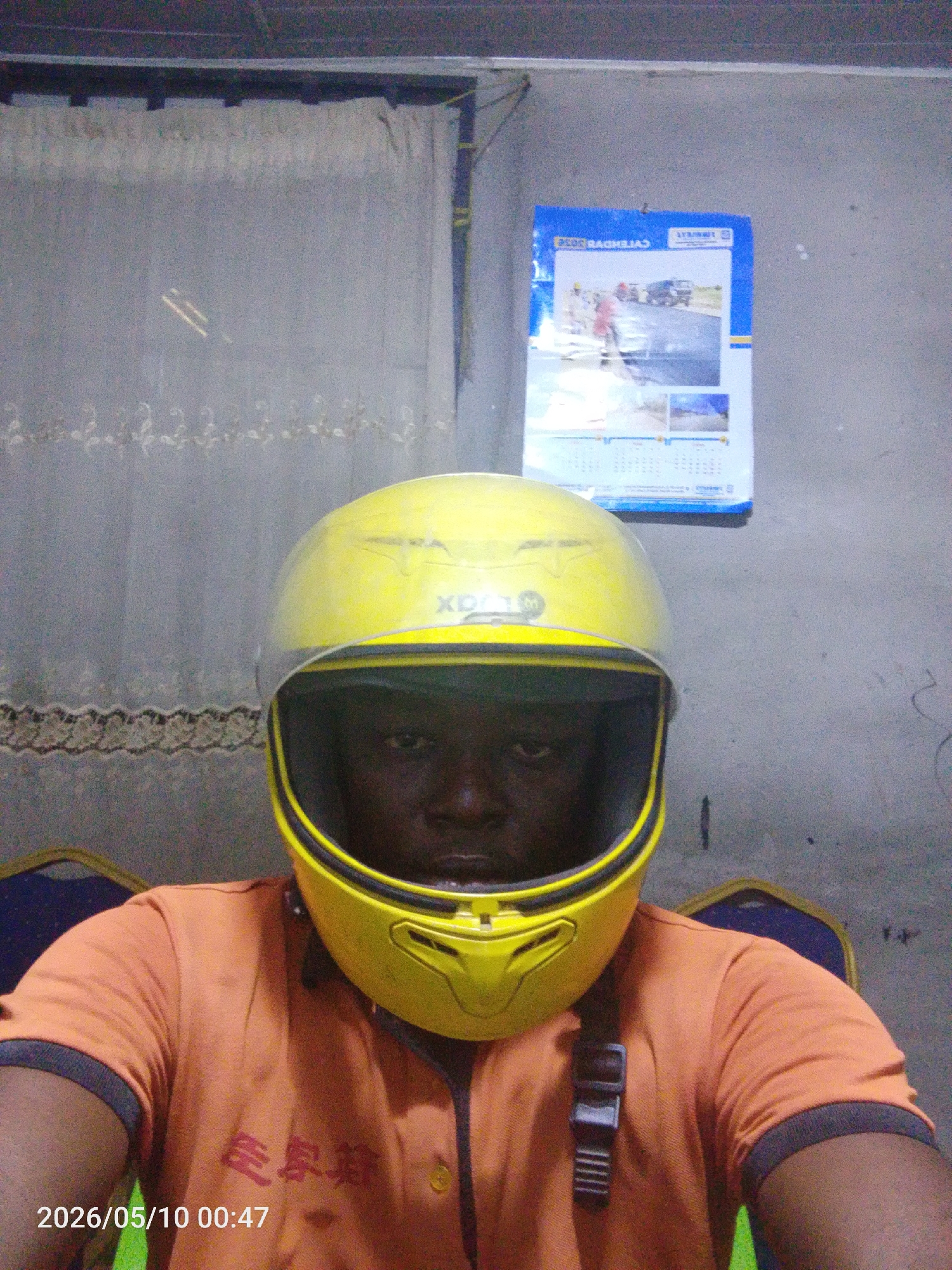 A person wearing a yellow helmet and an orange shirt with East Asian characters printed on it is seated. In the background is a gray wall with a calendar hanging on it and a gray lace curtain. The scene appears to be indoors. The location is implied to be Lokoja, Nigeria. The date and time "2026/05/10 00:47" are displayed in the lower-left corner.