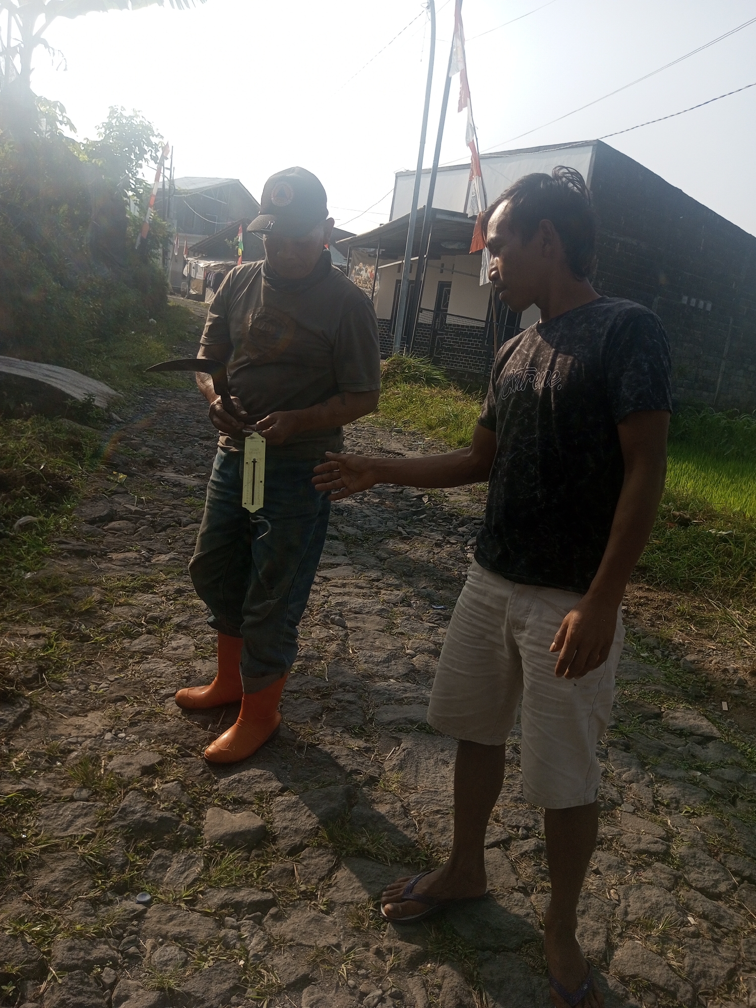 Two individuals are standing on a cobblestone path in what appears to be a rural or semi-rural area. The man on the left, wearing a dark t-shirt, jeans, and orange boots, holds a tool resembling a sickle and a hanging scale. The man on the right, wearing a patterned t-shirt and light-colored shorts, is gesturing with his right hand towards the other man. The background shows buildings, some with flags, and a grassy field. The scene is outdoors and appears to be during daylight hours. The location is identified as Krasak, Indonesia.