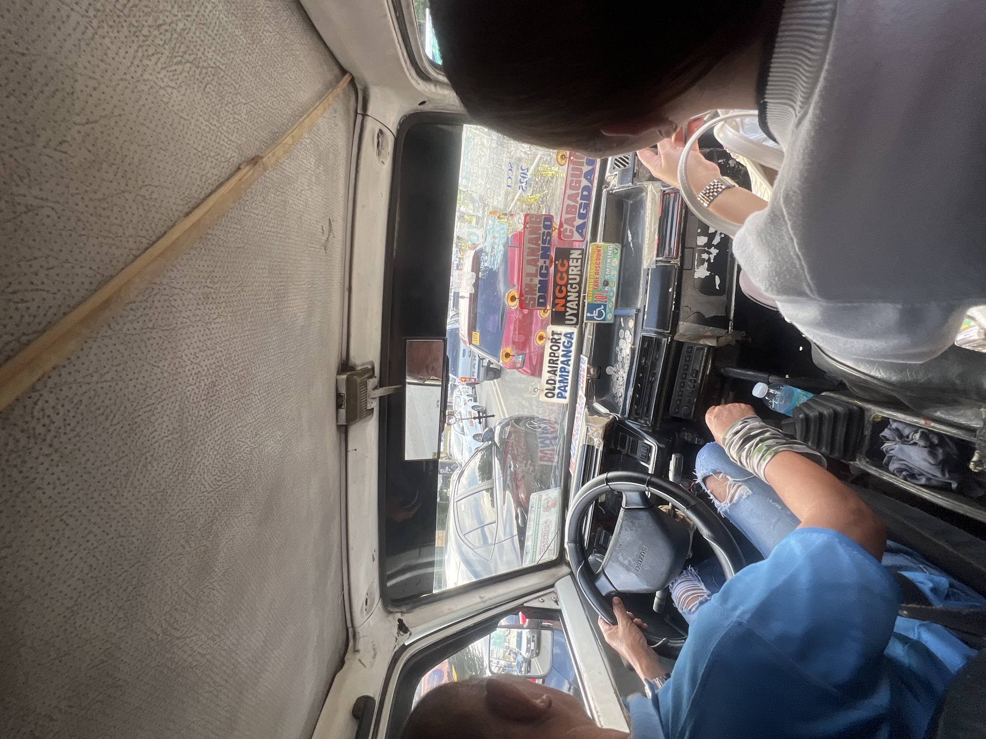 The image shows the interior of a public utility vehicle in Davao City, Philippines, presumably a jeepney, with two people visible. The driver, wearing a blue shirt, is holding the steering wheel with his left hand and operating the gearstick with his right hand. His left wrist has a watch and his right wrist is covered in a camouflage sleeve. He is wearing torn blue jeans. A passenger, wearing a white sweater, is partially visible in the upper right portion of the frame.
The vehicle's dashboard is cluttered with various stickers, route signage, and regulatory notices typical of jeepneys in the Philippines. The windshield is also covered with stickers, partially obscuring the view of traffic and the urban environment outside. Prominent stickers include destination routes like "OLD AIRPORT PAMPANGA", "UYANGUREN", "CABAGUI", "SM-LANANG", "DMC-NSO", "NCCC", and "AGDA". Signage includes an image of a person in a wheelchair. The interior is visibly aged and shows wear.