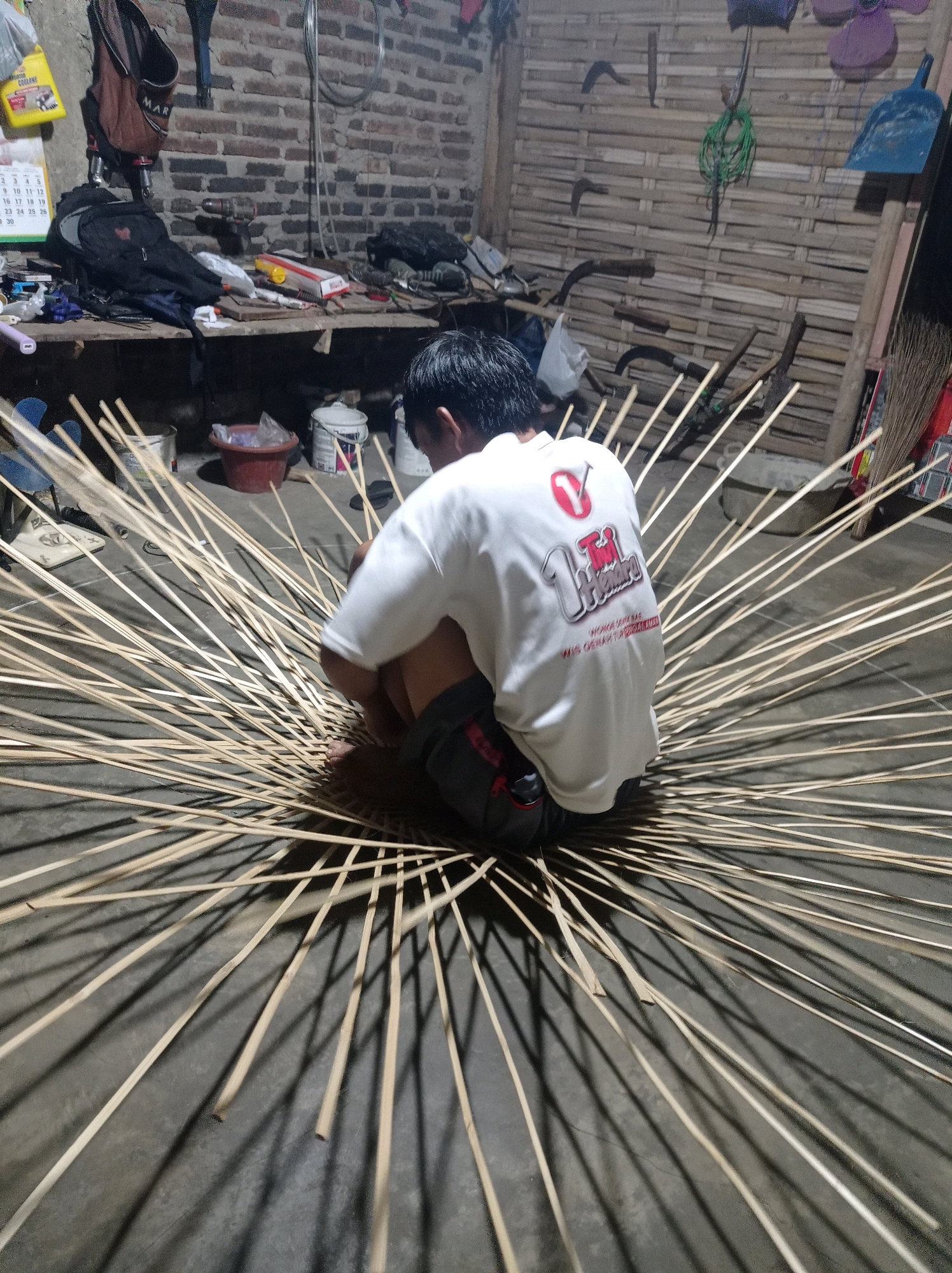 In Kedungbenda, Indonesia, a person is seen crouching on the floor of a workshop, surrounded by numerous thin wooden poles radiating outwards. The person is wearing a white t-shirt with red and white lettering on the back and dark shorts. They appear to be in the process of weaving or constructing something with the poles, possibly an umbrella or a fan, as the poles are arranged in a circular pattern. The workshop environment is rustic, with exposed brick walls, wooden shelves containing tools and various items, and bamboo-paneled walls in the background. Sunlight casts long shadows from the poles onto the floor, indicating the presence of a light source, likely a window or lamp, from the left side of the frame. Various tools and household items are visible on the shelves, suggesting a working space dedicated to crafts or repairs.