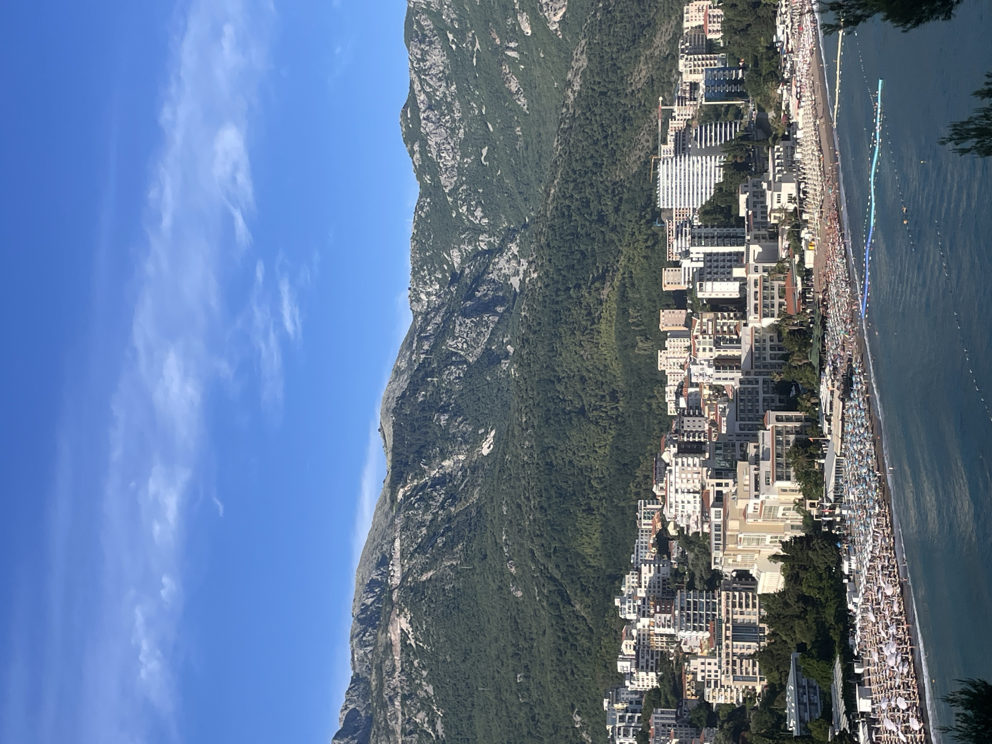 This image captures a panoramic view of the city of Boreti, Montenegro, nestled at the foot of a lush green mountain range under a clear blue sky with scattered white clouds. The foreground features a bustling beach with rows upon rows of white umbrellas and lounge chairs, indicating a popular tourist destination. The turquoise water of the Adriatic Sea gently laps at the shore, with some swimmers and buoys visible in the water. Behind the beach, a dense urban landscape of multi-story buildings, predominantly white and beige, extends inland. These buildings range from modern hotels to apartment complexes, showcasing a mix of architectural styles. The mountains, covered in dense green foliage and rocky outcrops, rise dramatically behind the city, forming a picturesque backdrop. The sunlight casts clear shadows, suggesting it is a bright, sunny day. The scene depicts a vibrant coastal resort town, alive with activity and bathed in natural light.