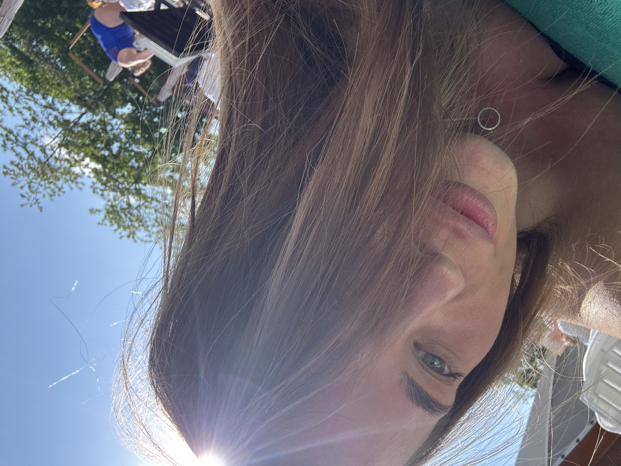 The image shows a close-up selfie of a person with light brown hair and green eyes. The person is wearing a circular earring. Bright sunlight illuminates the lower portion of their face. In the background, partly obscured by the person's hair and sunlight, are trees, a blue sky, and people seated at tables. Based on the location request, this scene is situated in Sosnovy, Russia.