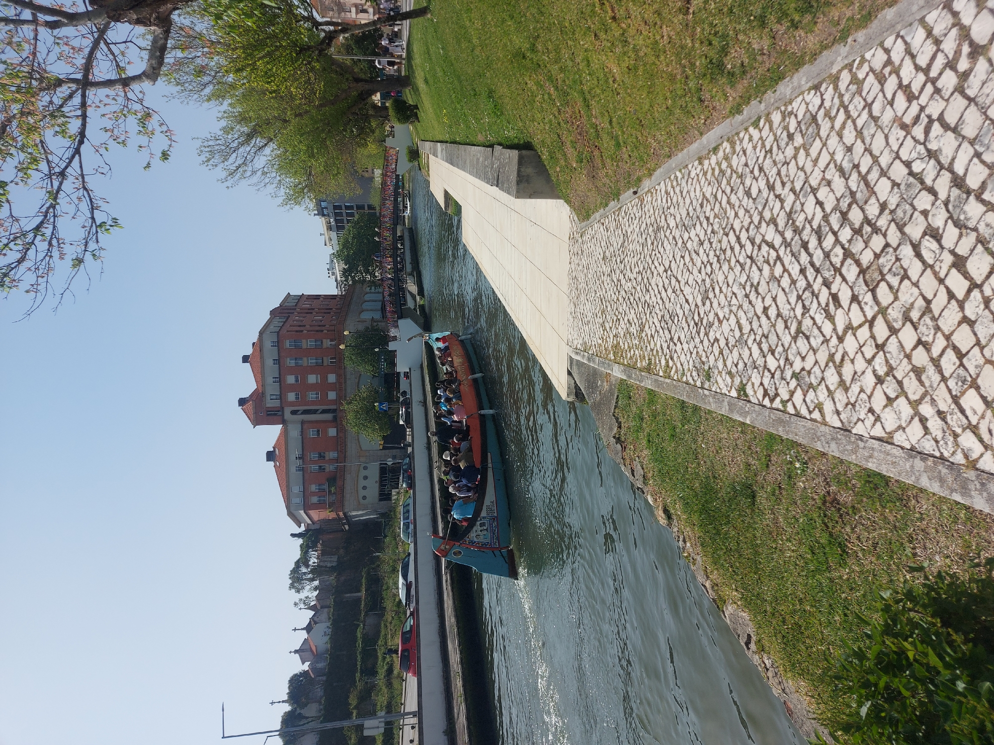 The image captures a canal scene in Aveiro, Portugal, featuring a moliceiro boat filled with tourists navigating the waterway. The boat is distinctively decorated, indicative of traditional Portuguese design. The canal runs between buildings and under bridges, with a view of the cityscape extending beyond the waterway. Cobblestone walkways and grassy banks line the canal, providing pedestrian access along the water's edge. The sky is clear, suggesting a sunny day.