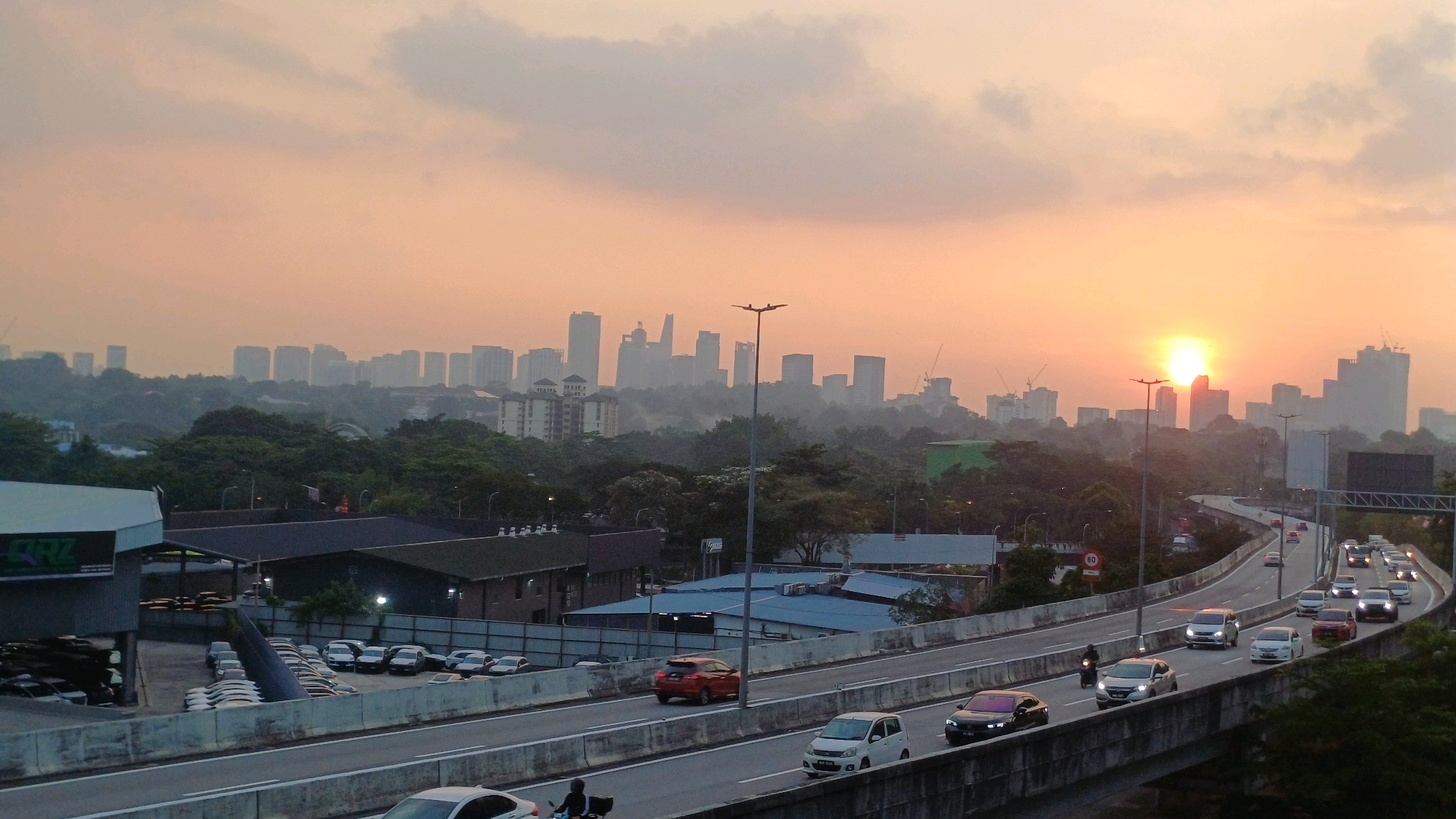 The image depicts a panoramic view of Kuala Lumpur, Malaysia, at sunset. The sky is a warm orange and yellow hue, indicating the setting sun, which is partially obscured by buildings on the horizon. A dense cityscape with numerous skyscrapers of varying heights stretches across the background, some appearing faded due to atmospheric haze. In the middle ground, a multi-lane elevated highway is visible, with cars and a motorcycle in motion. Trees and some industrial buildings with corrugated roofs are situated between the highway and the distant city skyline. The foreground shows a section of the highway and the edge of a building with the logo "QRZ" and stacked tires. The overall scene captures a typical urban environment during rush hour as daylight fades.