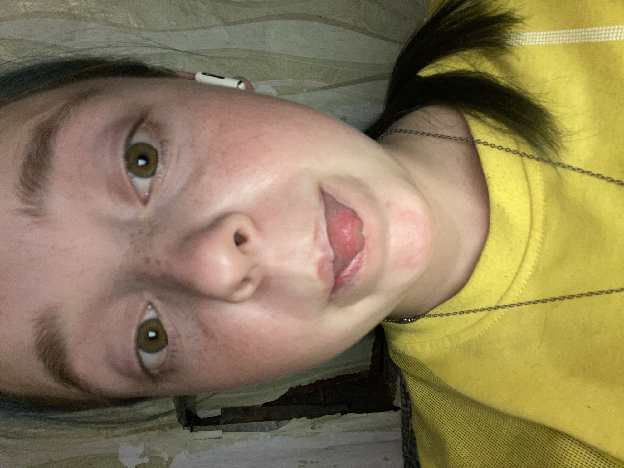 This is a close-up, frontal, eye-level selfie of a person with fair skin and freckles. The person has brown eyes and is wearing a yellow shirt and a silver chain necklace. An AirPod is visible in their right ear. The background appears to be a wall with peeling wallpaper, suggesting an indoor setting. The image does not provide any information about the location being in Nova Praha, Ukraine.