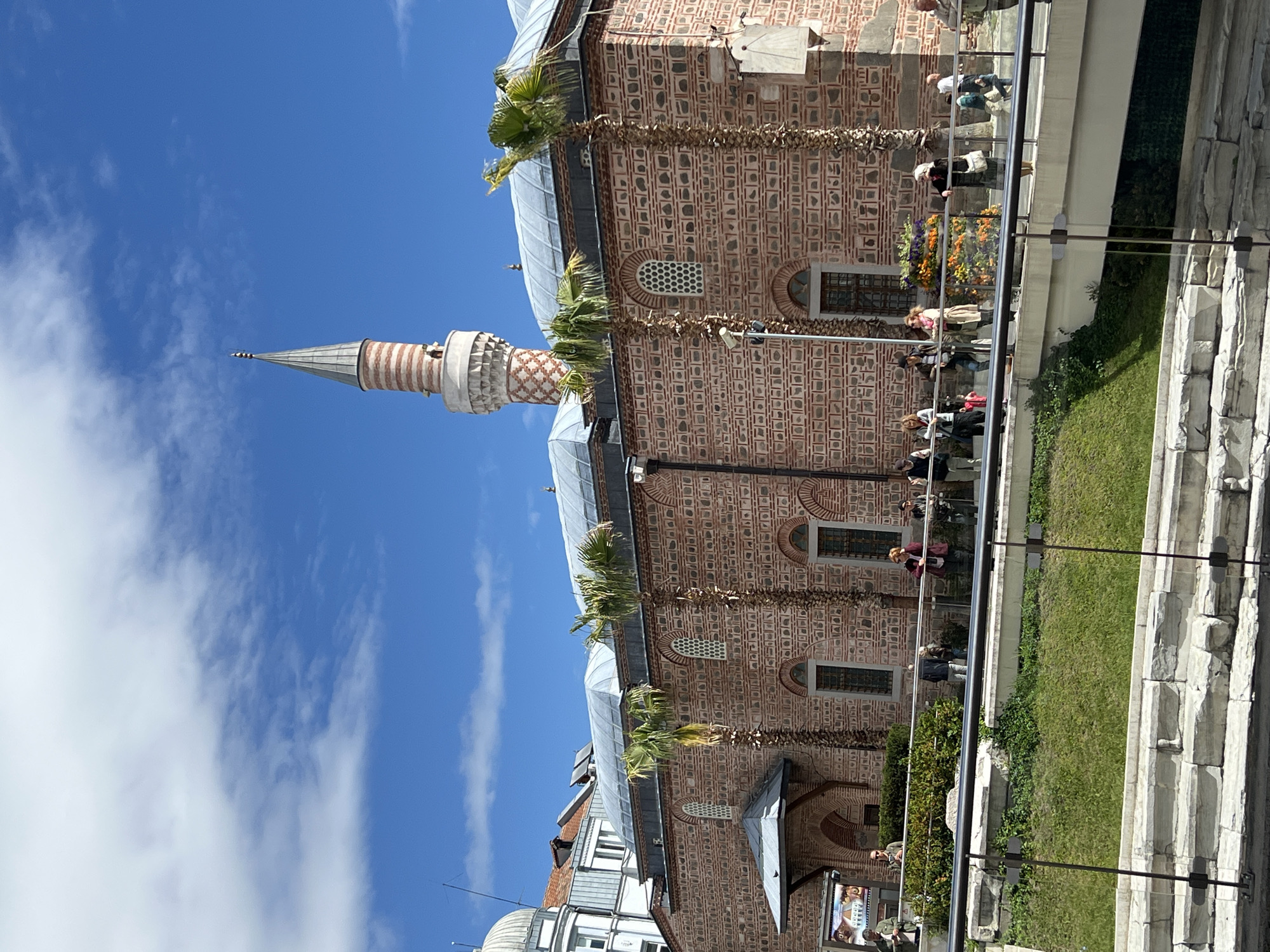 The image shows the Dzhumaya Mosque in Plovdiv, Bulgaria. The mosque is brick-built with a light-colored tower with a conical spire rising into a blue, partly cloudy sky. Palm trees grow along the mosque's exterior. People are visible on the mosque grounds. The mosque is partially reflected in a glass barrier that overlooks a grassy and landscaped area. A stone structure is also visible on the right side of the image. Other buildings are partially visible behind the mosque.