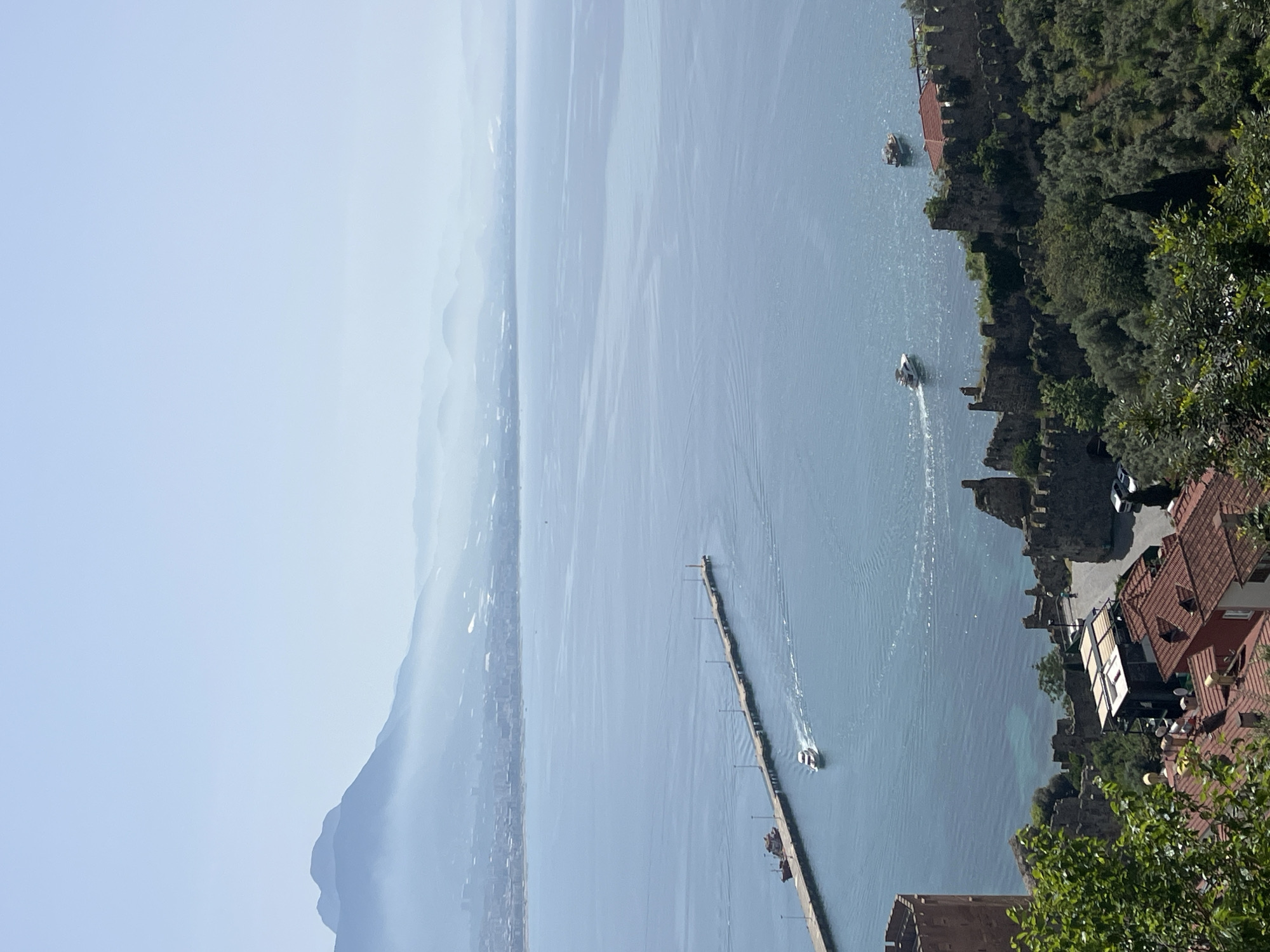 The image depicts a daytime aerial view of the coastline and sea in Alanya, Turkey. The shot appears to have been taken from an elevated position, looking down towards the water. A long pier stretches out into the sea, with several boats visible both near the pier and further out in the water.
Along the right side of the image, a section of the old city wall runs along the coastline. Buildings with red-tiled roofs are visible among the trees and fortifications. A few cars are parked on a road next to the buildings. The left side of the image shows the coastline stretching into the distance, with mountains visible in the background. The sky is a hazy blue, indicating a clear but possibly humid day.