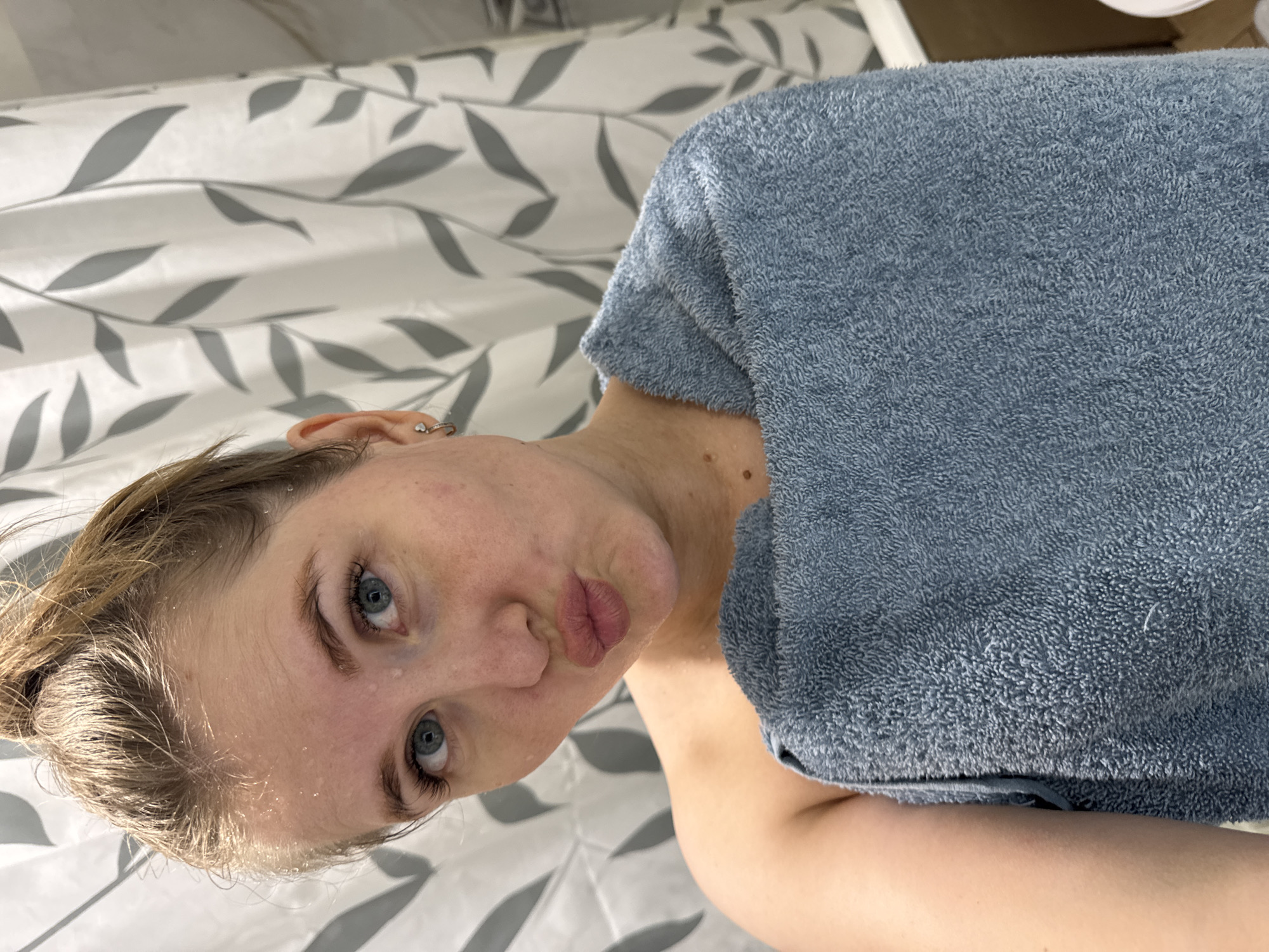 A young woman with wet blonde hair and blue eyes poses in a bathroom. Her face is angled towards the camera, and she is puckering her lips. A blue towel is draped over her shoulder. The background includes a shower curtain with a gray leaf pattern. The location is a bathroom, but the city Ферма, Russia, is not discernible from the image.