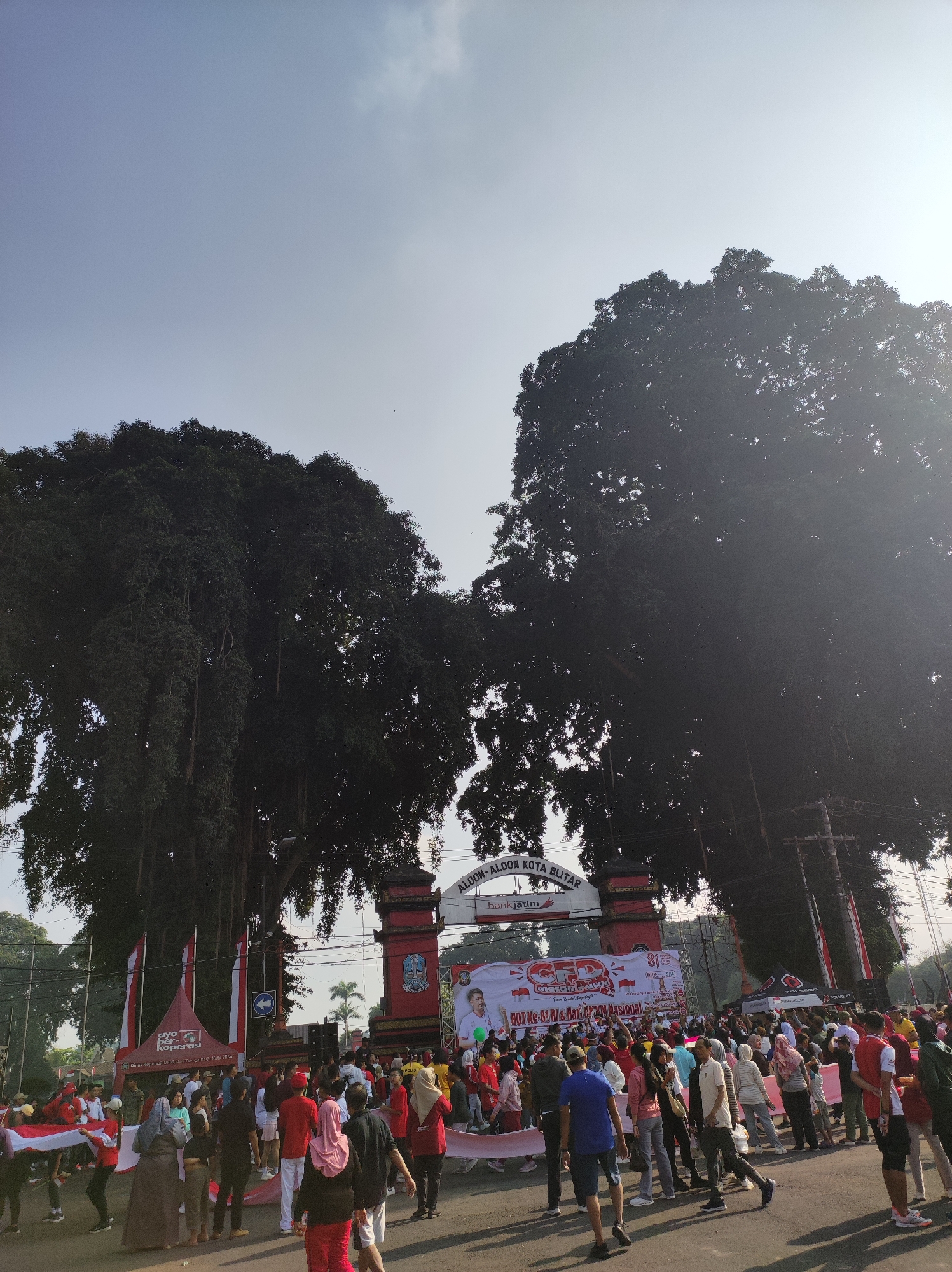 In the city of Blitar, Indonesia, a large crowd gathers in an open area with a prominent archway at its center. Two colossal banyan trees with cascading aerial roots flank the archway, their dense foliage creating a dramatic silhouette against the bright sky. The archway itself is red with ornate details and features banners and flags. One large banner reads "ALUN-ALUN KOTA BLITAR," identifying the location as Blitar City Square. Another banner displays the text "HUT RI Ke-81" and "MEMPERKOKOH PERSATUAN BANGSA" along with images of people and text in Indonesian. Numerous people, dressed in casual attire, are visible in the foreground and midground, many wearing red and white, suggesting participation in a national celebration or event. Some individuals are holding or unfurling a large Indonesian flag. The scene appears to be a public gathering or event, possibly a parade or festival, taking place outdoors during the day.