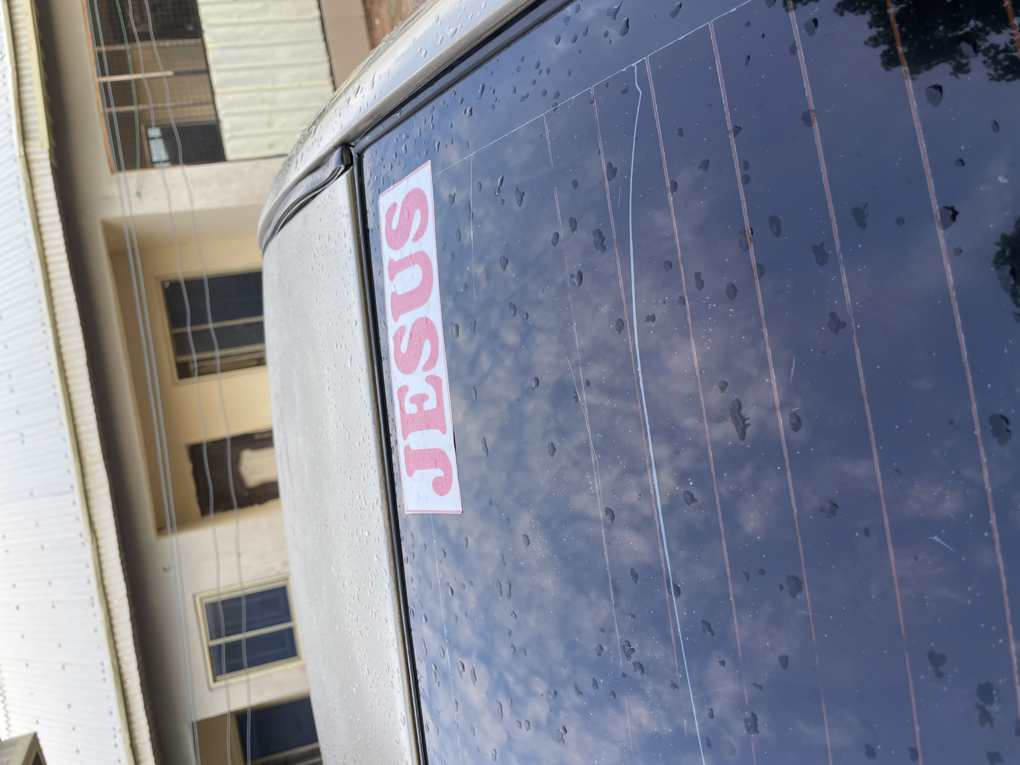 The image depicts the rear exterior of a light-colored vehicle, likely a sedan, parked in the city of Jalingo, Nigeria. The car's roof and rear window are covered in numerous small water droplets, indicating a wet surface.
On the upper left interior of the car's dark-tinted rear window, a rectangular sticker with the word "JESUS" is prominently displayed. The letters are red and uppercase, set against a white background. Multiple thin, horizontal defroster lines are visible across the rear window.
Reflected in the car's rear window and visible in the background is a multi-story building with a light-colored (beige/cream) facade and a light gray corrugated metal roof. The building features dark, recessed openings, some appearing to be windows with vertical mullions, and others resembling doorways or unfinished sections. Several thin utility lines, possibly electrical or communication cables, traverse horizontally across the midground in front of the building. The reflection in the car's window also shows a cloudy sky and dark foliage from what appears to be a tree or bush on the lower right side. No individuals are present in the scene.