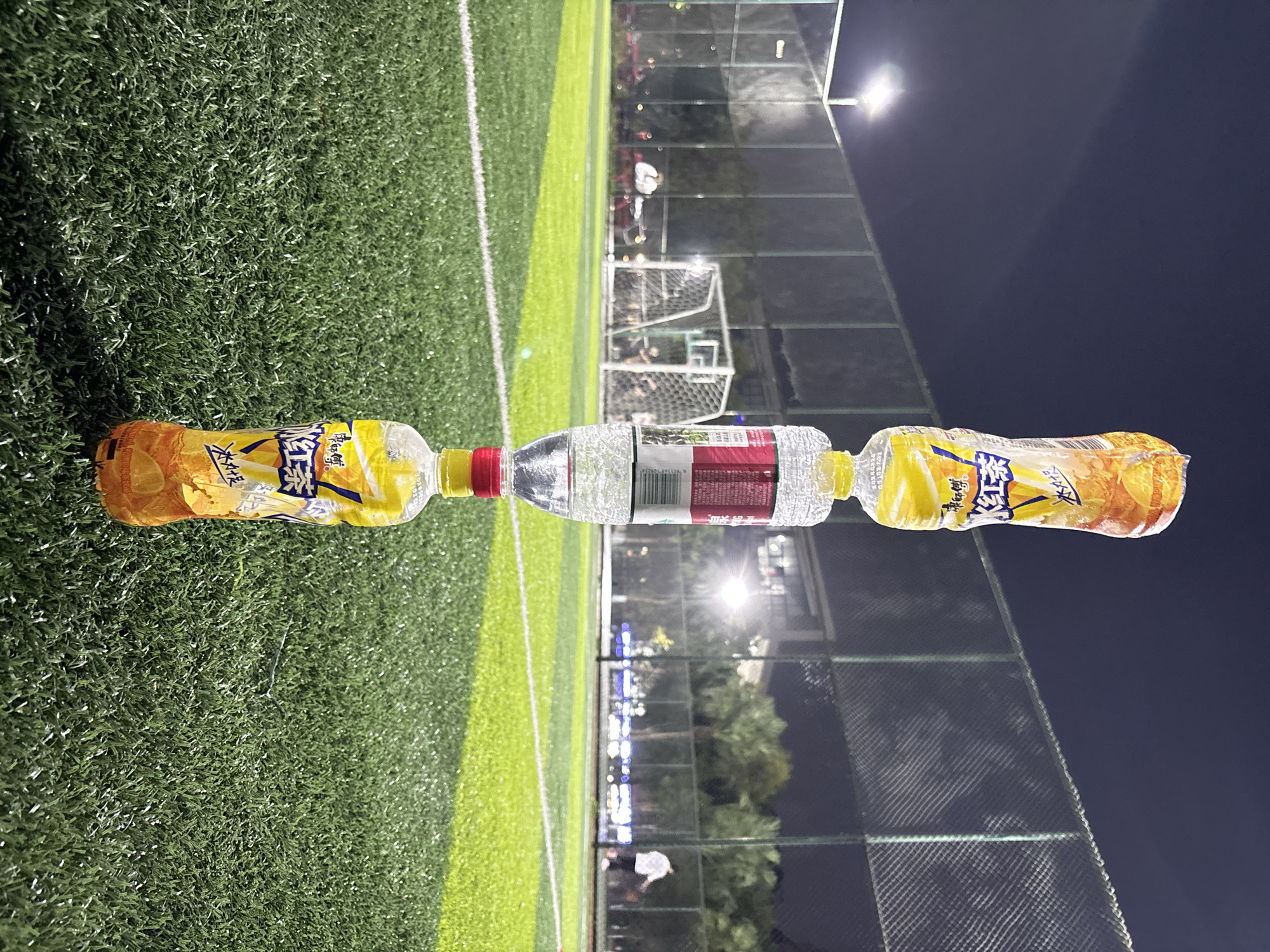 The image depicts three bottles horizontally connected on a green field. Two bottles on either end are of the same brand of iced black tea, while the bottle in the middle is filled with clear water with a red label. In the background is a soccer field surrounded by netting. Overhead lights illuminate the scene. The location is in the city of Jiangjin, China.