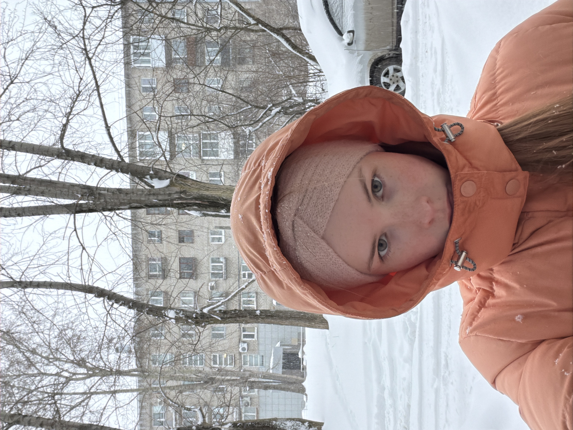 This is a portrait-oriented photograph captured outdoors in a winter setting in Berezniki, Russia. The main subject is a young person, likely female, positioned in the right foreground, looking directly at the camera with a neutral expression. They are wearing a peach or light orange colored hooded winter jacket, and a light pink knitted headband or hat is visible under the hood. Small specks of snow or ice are present on their jacket and headband.
The ground is covered in a thick layer of white snow, visible in the lower and right parts of the image. Prominent bare tree branches, some with traces of snow, extend across the left side and mid-ground, partially framing the background. In the background, on the left, stands a multi-story residential building with a light-colored facade and multiple windows. A white car, heavily covered in snow, is partially visible in the upper-right background, behind the person. The scene conveys a cold, snowy, daytime atmosphere, suggesting either active snowfall or recent heavy snow. There is no visible text in the image.