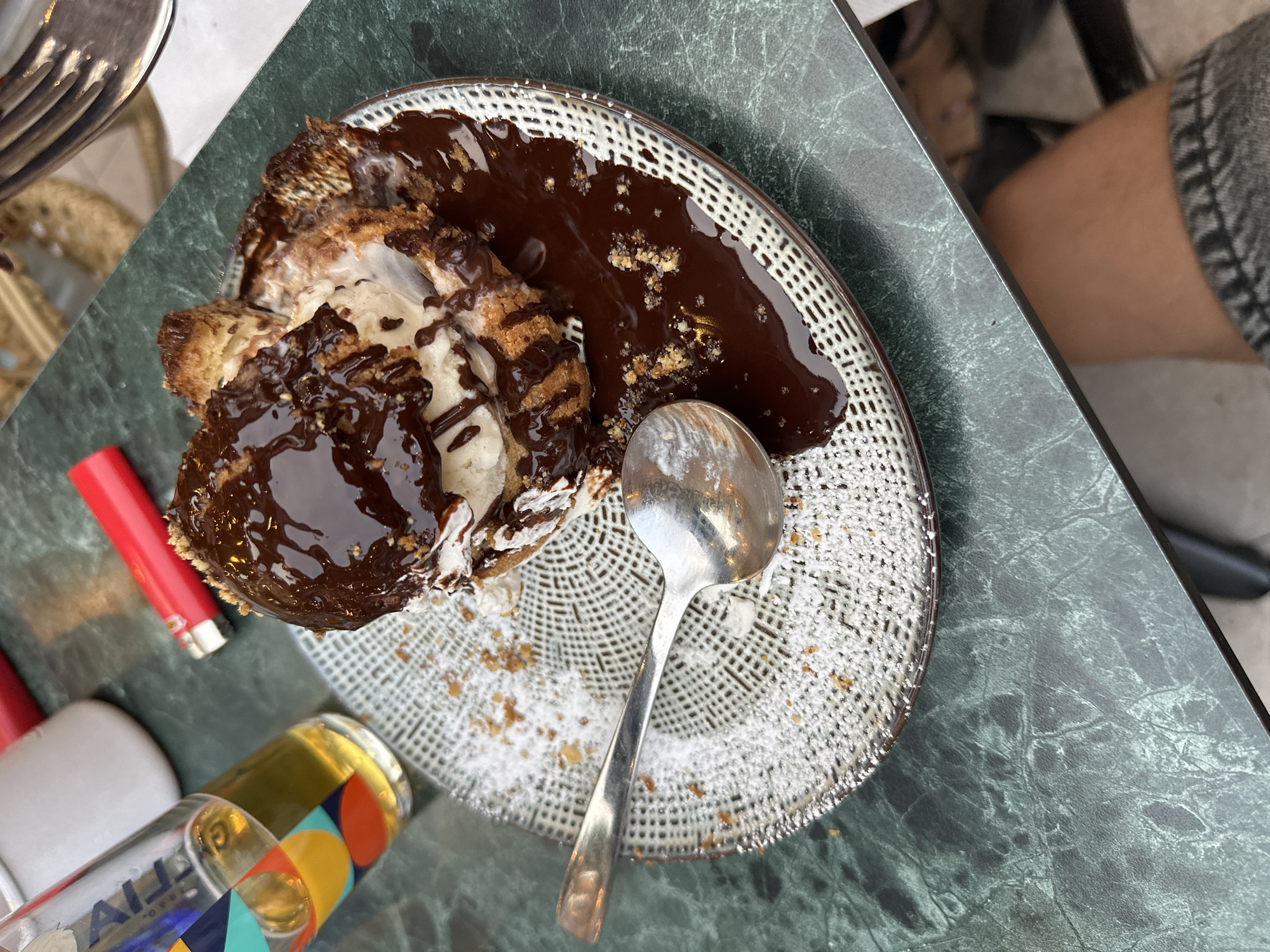 A close-up, overhead shot shows a dessert on a dark green, marble-patterned table. The dessert, likely a fried pastry filled with ice cream and drizzled with chocolate sauce, sits on a decorative gray and white patterned plate. A silver spoon rests on the plate, partially covered in powdered sugar. To the left of the dessert, a glass of amber liquid, possibly beer, is visible. A red lighter is positioned behind the glass. The background is slightly blurred, with hints of other objects and a person's leg and knee visible in the bottom right corner. The lighting suggests an outdoor or well-lit indoor setting.