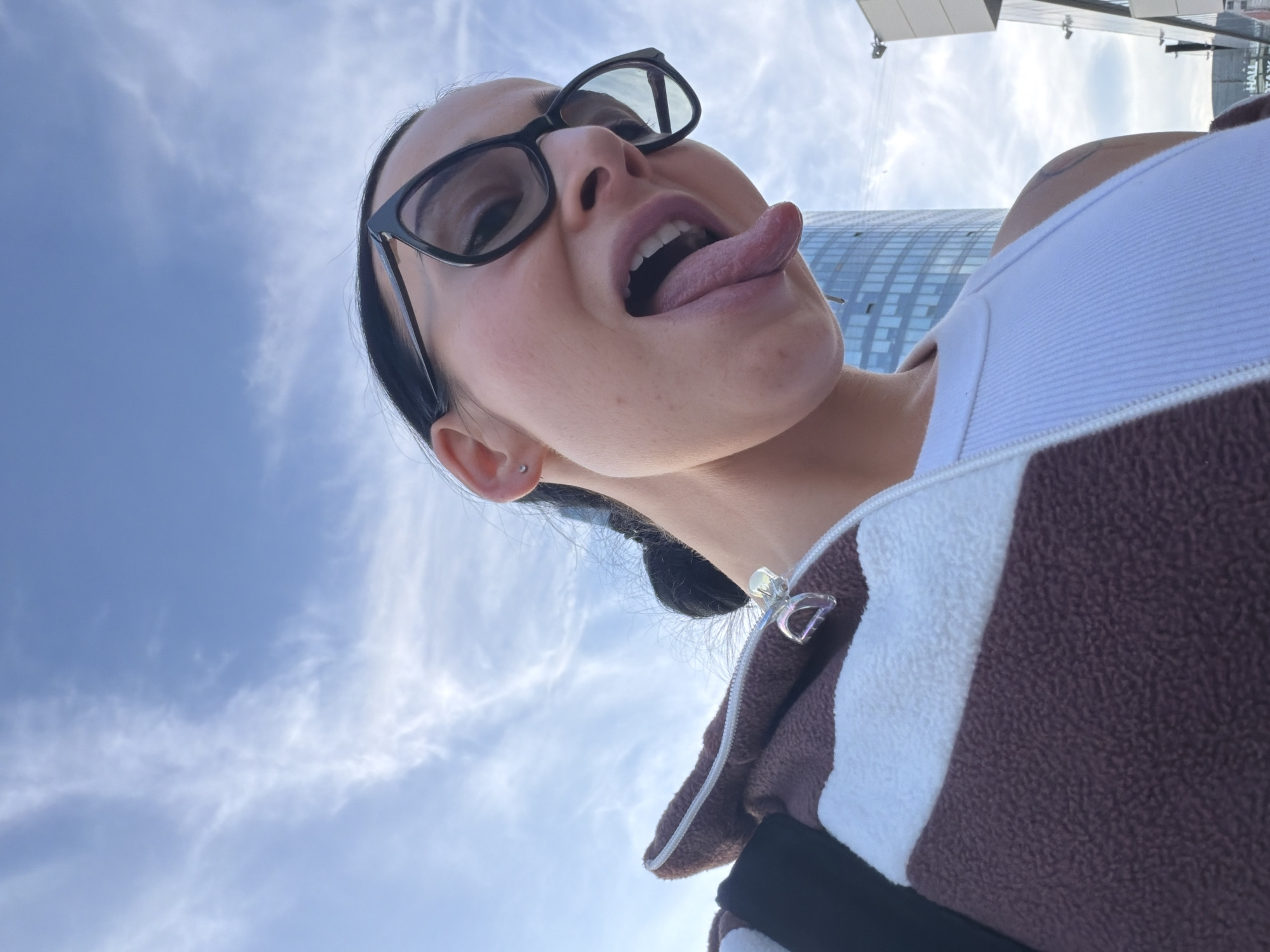 A low-angle, upward-facing shot captures a person looking directly at the camera, making a playful expression by sticking out their tongue. The individual wears black-framed glasses, a white ribbed tank top, and a brown and white striped fleece jacket with a zippered front. A clear, possibly iridescent, zipper pull is visible on the zipper. The person has a small stud piercing in their left earlobe and dark hair pulled back.
The background features a bright blue sky with scattered white, wispy clouds, indicating daytime. Behind the person's right shoulder, a tall, modern building with a facade of numerous windows is visible, reflecting the sky. To the upper right, another section of a building or structure is seen, displaying faint text. The visible text appears to be "HALL" and below it, "TAXI" (likely written in Cyrillic, which aligns with the Saint Petersburg, Russia context).
The image conveys a lighthearted and candid moment, possibly a selfie, with the urban environment serving as a backdrop under clear skies.