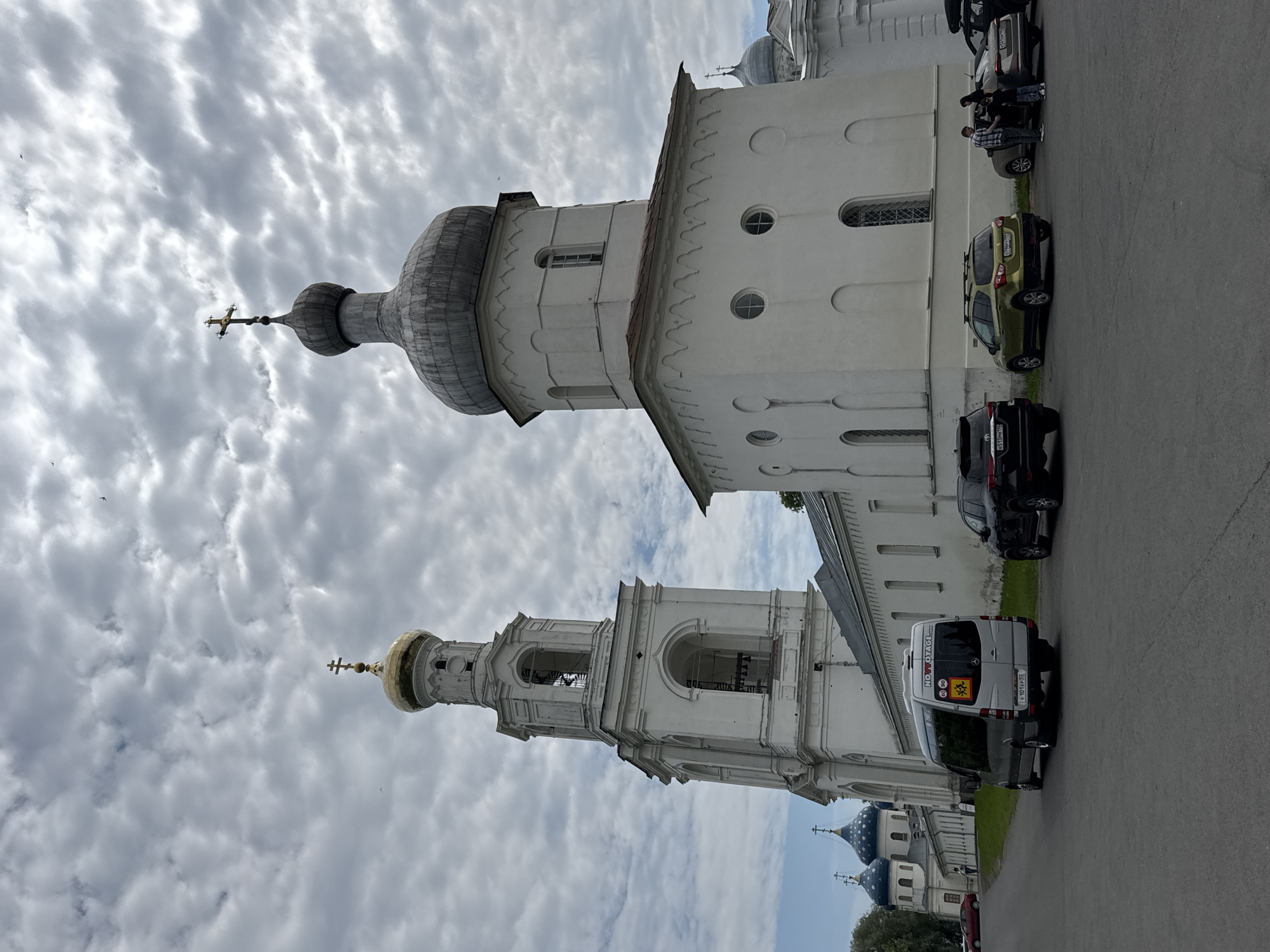 The image depicts the Bell Tower and Assumption Cathedral of the Saint Sophia Monastery in Veliky Novgorod, Russia, under a cloudy sky. In the foreground, a paved area hosts several parked cars, including a silver Mercedes-Benz Sprinter van with the text "NOVOYAGE" and school bus icons visible on its rear. To the right of the van are a black SUV and a gold hatchback. Further right, two individuals are interacting near a dark sedan. The Bell Tower, a tall, white, multi-tiered structure with arched openings and a golden dome topped with a cross, stands prominently on the left. Adjacent to it, a smaller white building with a domed roof, also topped with a cross, is visible. In the background, partially obscured by the main structures, are blue-domed churches with crosses. The overall environment appears to be a historical and religious complex, likely a tourist destination, given the presence of vehicles and people.