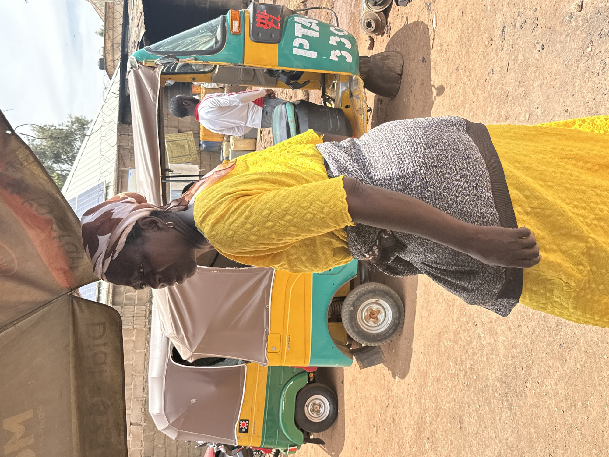 In Gura Top, Nigeria, an elderly woman wearing a yellow dress and a patterned headscarf stands in front of several auto rickshaws, also known as tuk-tuks. The vehicles are green and yellow. One has the number "330" written on it. A man in a white shirt is seated in one of the rickshaws. The ground is light brown and dusty. An umbrella partially obscures the foreground.