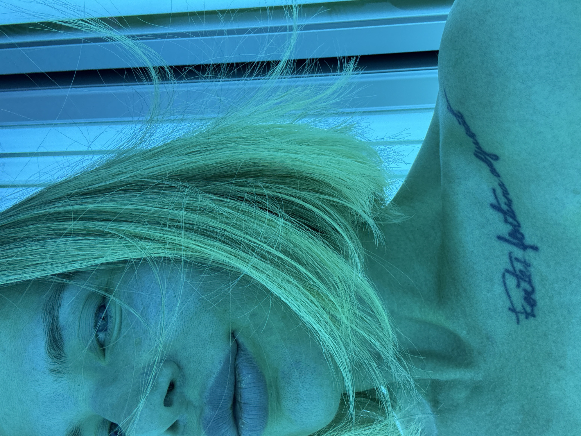 A close-up, angled shot depicts a fair-skinned individual with blonde hair, possibly female, lying beneath what appears to be a tanning bed. A tattoo is visible on the upper arm. The environment is illuminated with a distinct blue light. The location, Gorno-Altaysk, Russia, is not evident within the image details.