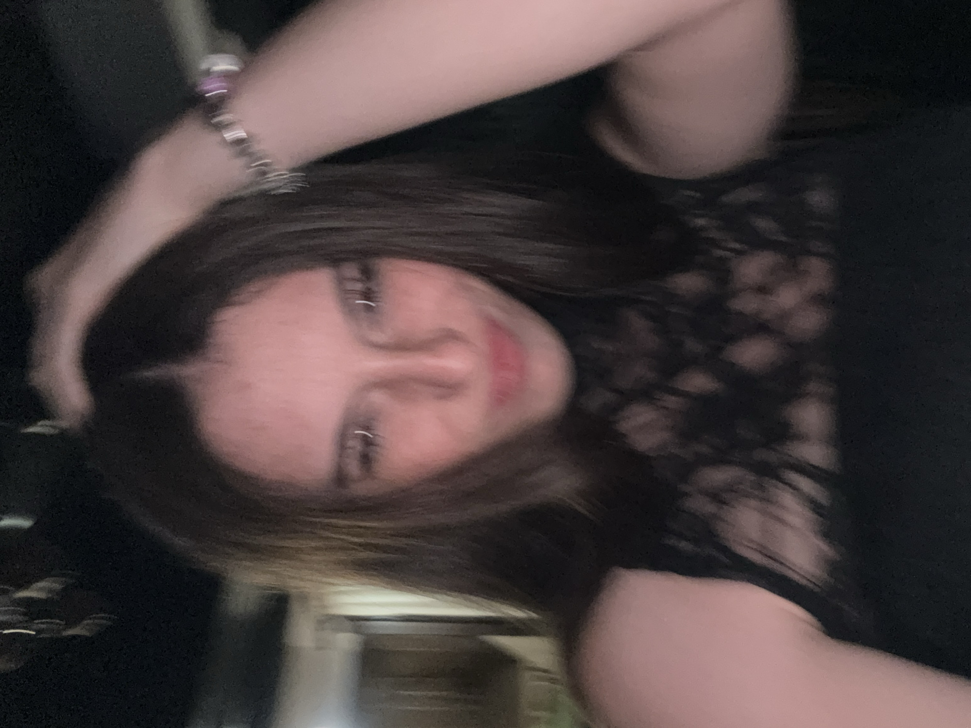 A young woman with long, dark hair is pictured in a dimly lit indoor environment. She is looking towards the camera with her hand raised to her head. She is wearing a black, lace-detailed top and a bracelet on her wrist. The background is out of focus, but appears to be a room with some furniture and possibly a doorway. There is no indication of the specific location in Tazha, Bulgaria.