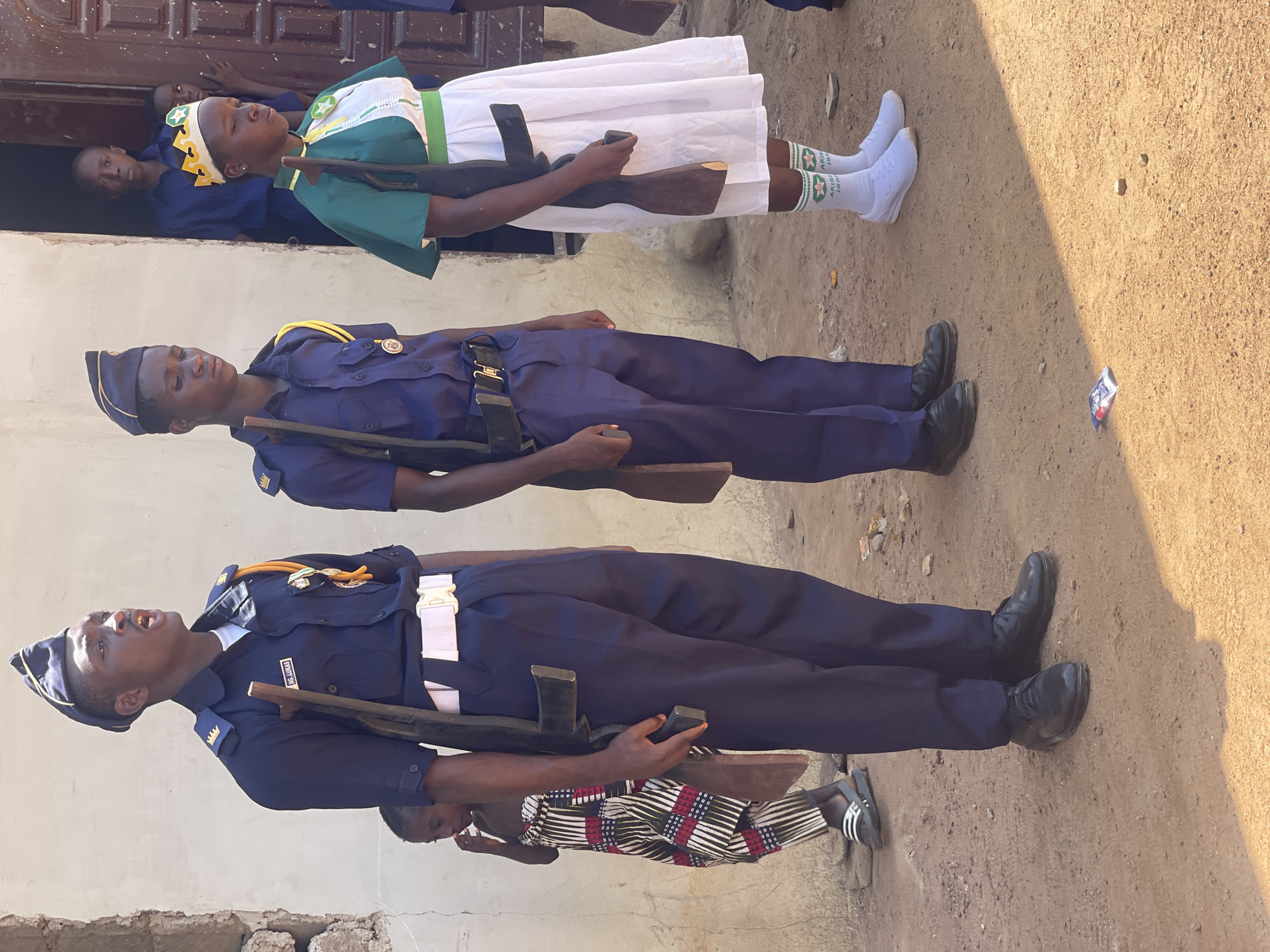 The image captures an outdoor scene in Andaha, Nigeria, depicting a formal gathering or parade involving several young individuals in uniform. The bright daylight and distinct shadows suggest it is daytime, with an unpaved, dusty ground serving as the setting.
In the foreground, two young males stand in formation, seemingly at attention. Both are dressed in navy blue uniforms with gold-colored epaulets, white belts, and matching garrison caps. They each hold a wooden mock rifle. The male closest to the viewer has a nametag on his uniform that reads "AWNIKI OIG". Behind the second male, a young child in a patterned outfit is partially visible.
Adjacent to them, a young female also stands formally. She wears a green and white uniform, comprising a green top with white trim, a white skirt, and a white headpiece with yellow geometric patterns. She has white socks adorned with green text that reads "ARISA IS" (the last word partially obscured) and white athletic shoes. She also holds a wooden mock rifle.
In the background, several other individuals, including children, are visible observing the scene from a doorway of a light-colored building with a dark wooden door. The disciplined postures of the uniformed individuals suggest they are part of a youth organization or cadet corps participating in a drill or ceremonial event.