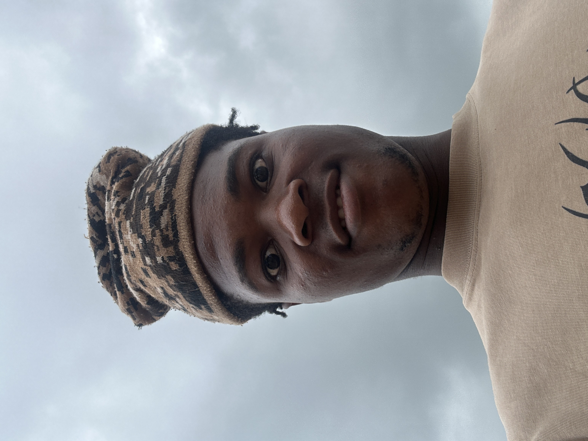 A close-up, low-angle selfie of a young Black person wearing a brown and black patterned beanie and a tan t-shirt. The person is looking up at the camera with a slight smile. Their hair is visible underneath the beanie, with a few strands sticking out. The background is a cloudy sky, suggesting an overcast day. The image appears to have been taken outdoors. The subject's face is the primary focus, with the beanie framing their forehead and the t-shirt visible on their shoulder. The overall impression is casual and informal, typical of a selfie. There is no visible text in the image. The location context of Ihiagwa, Nigeria, might suggest a warm climate, and the cloudy sky could indicate the possibility of rain or a generally humid atmosphere common in tropical regions. The time of day is not definitively discernible from the lighting, but the brightness suggests it is daytime.