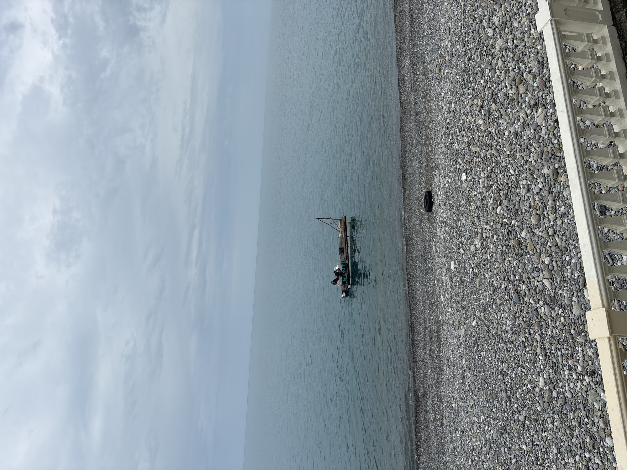 The image shows a calm, grey seascape. A small fishing boat with a crane-like structure is anchored in the water close to the shore. The shoreline is a pebble beach. A tire is laying on the beach. The beach is bordered by a decorative white concrete wall. The sky is overcast. Based on the provided crops, the location is in Sochi, Russia. No people are visible.