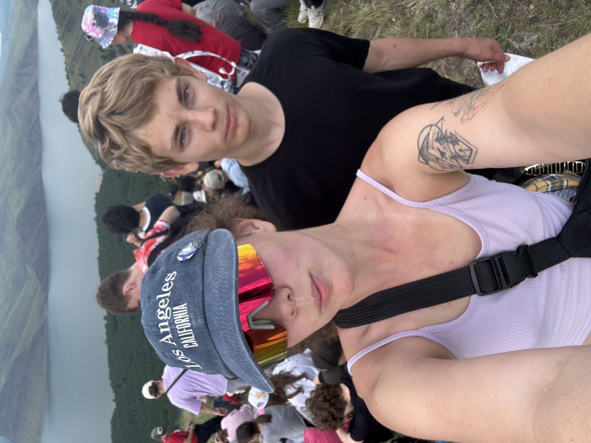 A medium shot shows a young woman in the foreground on the left, taking a selfie with a young man in the foreground on the right. She is wearing a light purple tank top, a black crossbody strap, and a blue baseball cap with "Los Angeles CALIFORNIA" embroidered on it. She is also wearing large, mirrored sunglasses with a rainbow-colored tint. The young man is wearing a black t-shirt. Both are looking towards the camera.
Behind them, a crowd of people are gathered on a grassy hillside overlooking a large body of water. The landscape in the background appears to be a mountainous region with a reservoir or lake. The water is calm and reflects the muted colors of the overcast sky. The vegetation on the hills is a mix of green grass and sparse trees. The overall atmosphere suggests an outdoor event or gathering in a scenic, possibly remote, location. The specific location is identified as Nikolo-Petrovka, Russia.