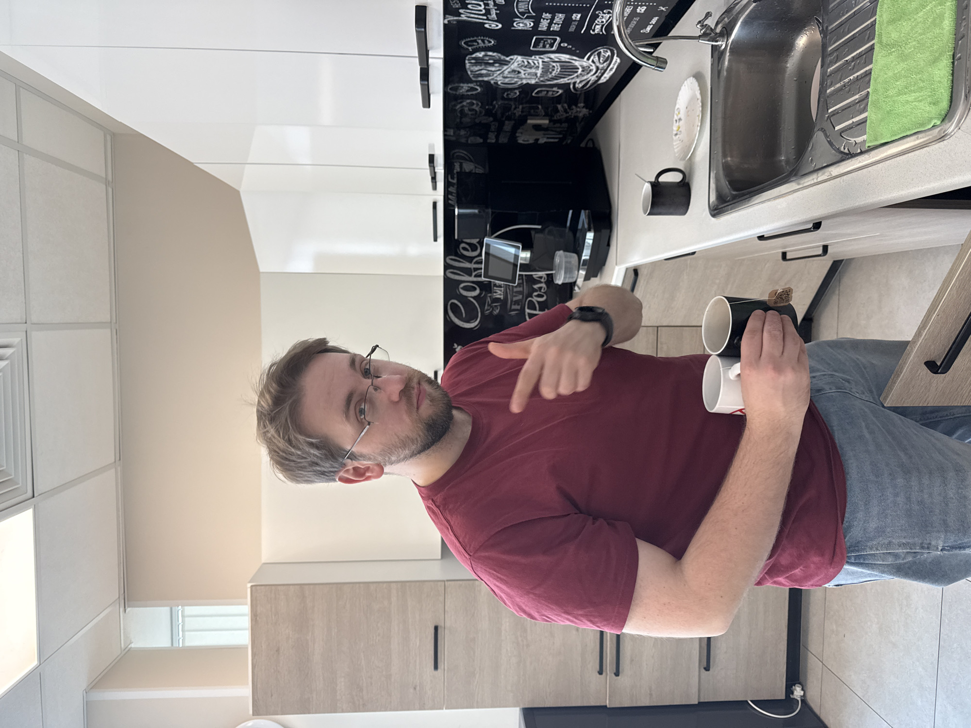 The image captures a young man in a modern kitchen or breakroom, possibly in Moscow, Russia. He is positioned in the foreground, facing slightly towards the viewer with his body angled, wearing a maroon short-sleeved t-shirt and blue jeans. He has light brown hair, a beard, and glasses, and holds a black and a white mug in his left hand, while his right hand points forward or to the mugs. A tea bag tag is visible on the black mug, suggesting he is preparing tea. His expression is neutral or slightly inquisitive.
The background reveals a well-equipped kitchen area. To his left, there are light wood-toned cabinets with black handles. To his right, a light-colored countertop features a stainless steel sink with a faucet, a drying rack, and a bright green dish towel. A white paper plate with a floral design and another black mug are also on the counter. A black automatic coffee machine stands prominently on the countertop. Above the counter are white cabinets, and a black backsplash patterned with white chalk-style coffee-themed illustrations and text. Visible words include "Coffee," "Latte," "Cappuccino," "Espresso," "Mocha," "Menu," "Have a great day," and "Since 2004," along with coffee cup and bean drawings. The ceiling above is composed of white panels. The overall setting appears to be a clean, contemporary interior, likely a daytime scene.