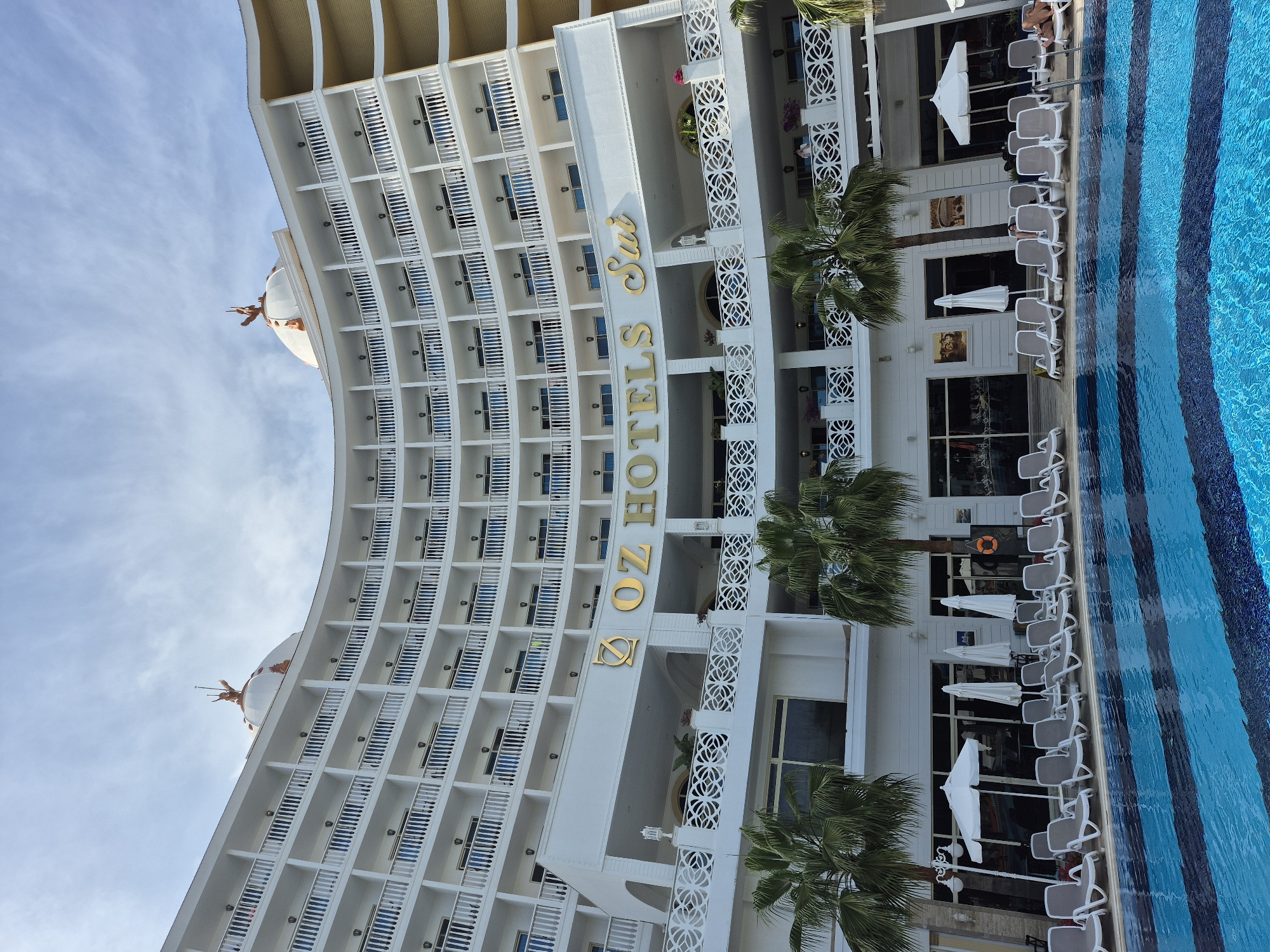 The image shows the exterior of the OZ Hotels Sui in Alanya, Turkey. The building is white with many balconies and the hotel's name displayed vertically in gold letters. Several palm trees are in the foreground. Adjacent to the building is a blue-and-white striped swimming pool surrounded by white lounge chairs and umbrellas. People are visible lounging by the pool. The sky is light blue with some clouds.