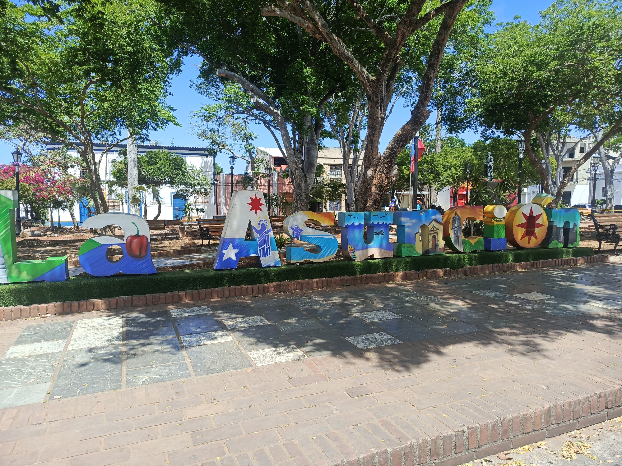 The image shows a sign spelling out "LA ASUNCION" in large, colorful letters in a plaza. The letters are raised on a brick platform lined with artificial turf. The background includes trees providing shade, buildings, and benches. The ground in the foreground is paved with brick and stone. The location is La Asuncion, Venezuela, indicated by the name on the sign. No people are clearly visible in the image. The scene appears to be a sunny day in a public park.