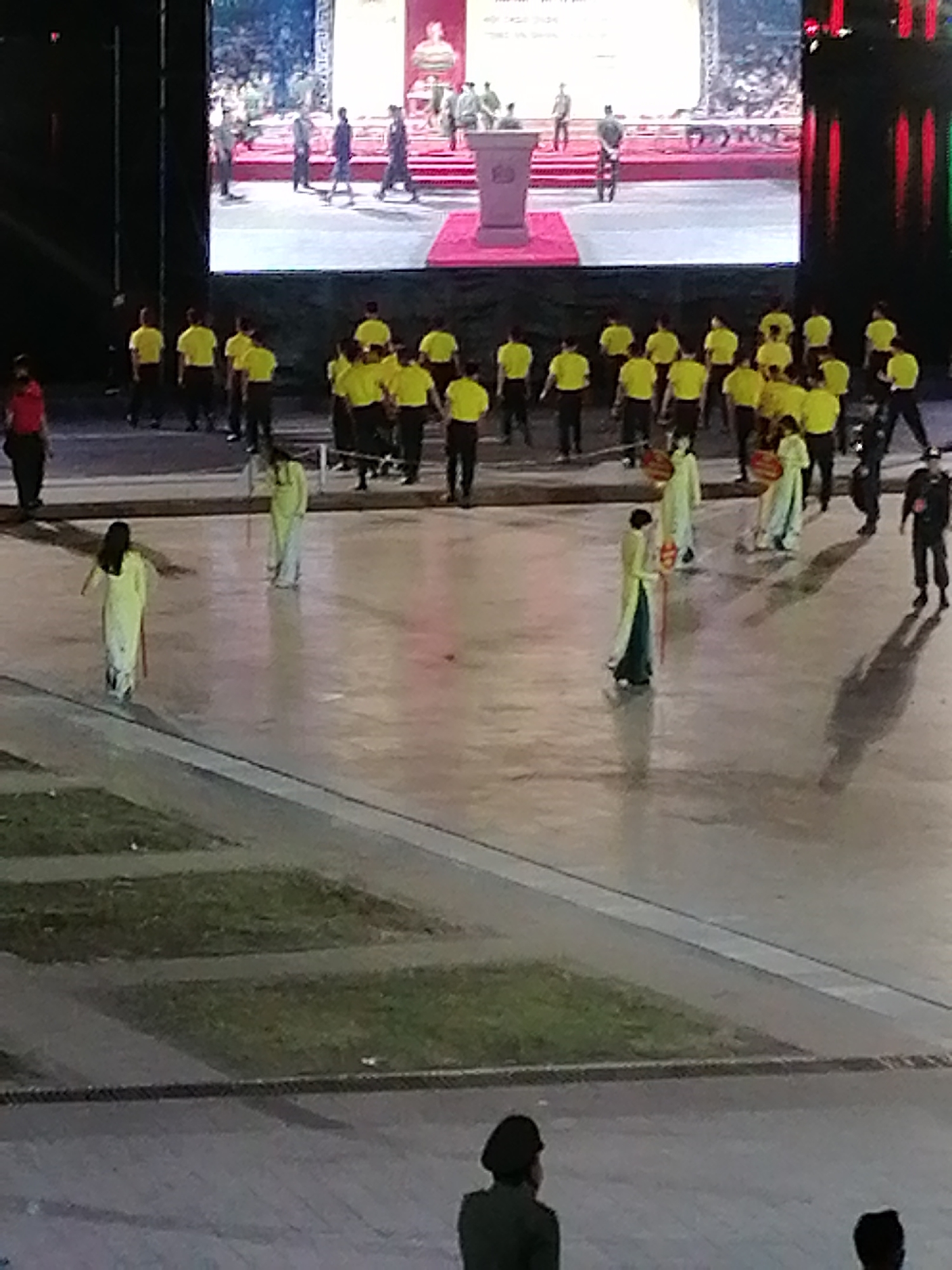 The image depicts an outdoor event, likely in Da Lat, Vietnam, given the architectural style visible on the large screen. The screen displays a performance or ceremony with people in historical or traditional attire on a raised platform. Below the screen, a large group of individuals in yellow shirts are gathered, some holding what appear to be poles or staffs. Further in the foreground, several people are dressed in long, flowing robes, possibly a traditional Vietnamese garment like an Ao Dai, some carrying decorative items. The ground is a smooth, reflective surface, possibly a plaza or paved area. In the very foreground, two individuals are visible from the back, one wearing a dark cap and uniform. The environment suggests a public gathering or festival.