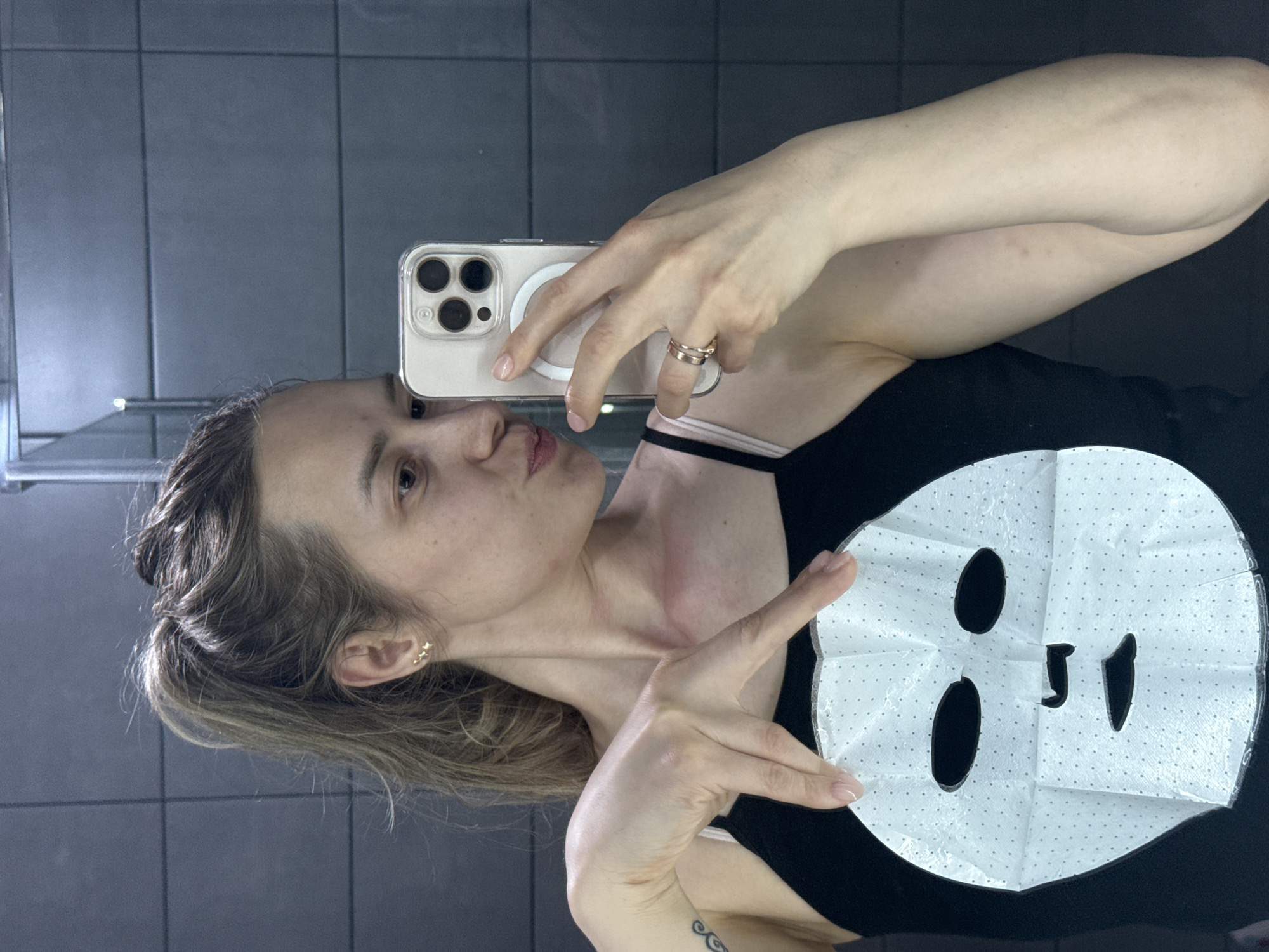 A person, likely a woman, is captured in a mirror selfie while holding a white sheet mask. She is looking at the camera with a playful expression, featuring puckered lips, and her head is slightly tilted.
She holds a light-colored smartphone with a clear case and a visible MagSafe ring in her left hand, featuring a distinct triple-lens camera module. In her right hand, she holds up the unfolded sheet mask, which is white with small black dots and cut-outs for eyes, nose, and mouth. She is making a "peace sign" gesture with her fingers on her right hand.
She is wearing a black tank top or camisole, with a lighter strap visible underneath. On her left ring finger, she wears multiple stacked rings, and her right ear features small, gold-colored earrings, possibly butterfly-shaped. A tattoo is visible on her inner right forearm. Her fingernails are painted with a light, neutral polish.
The background shows dark grey or black tiled walls, indicative of an indoor setting, possibly a bathroom or changing room. A metallic railing is visible in the upper left of the reflection. The lighting is even. The scene takes place in Chaikovsky, Russia. No visible text is present in the image.