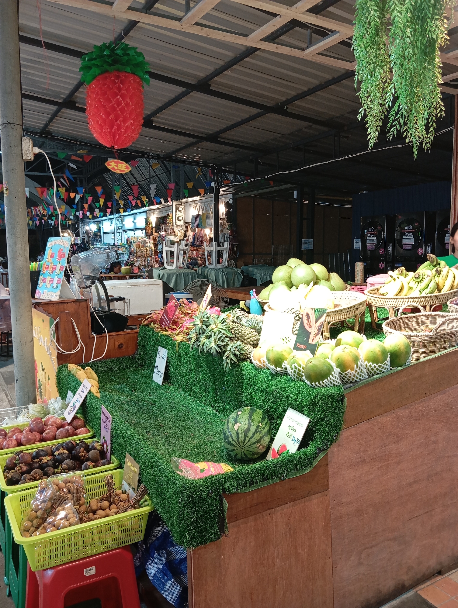 A tropical fruit stand is shown at a Karon, Thailand market. The stand is adorned with a large red and green pineapple decoration hanging from the ceiling. Colorful triangular flags are strung across the roof of the market. A variety of fruits, including papayas, bananas, watermelons, mangosteens, and longans, are displayed on green artificial grass. Some fruits are in baskets, while others are individually wrapped in netting. Signs with Thai script indicate the prices and names of the fruits. Behind the fruit stand, there are rows of what appear to be washing machines. The market is bustling with activity, with people browsing the stalls and purchasing produce. The overall atmosphere is vibrant and lively, characteristic of a Thai street market.