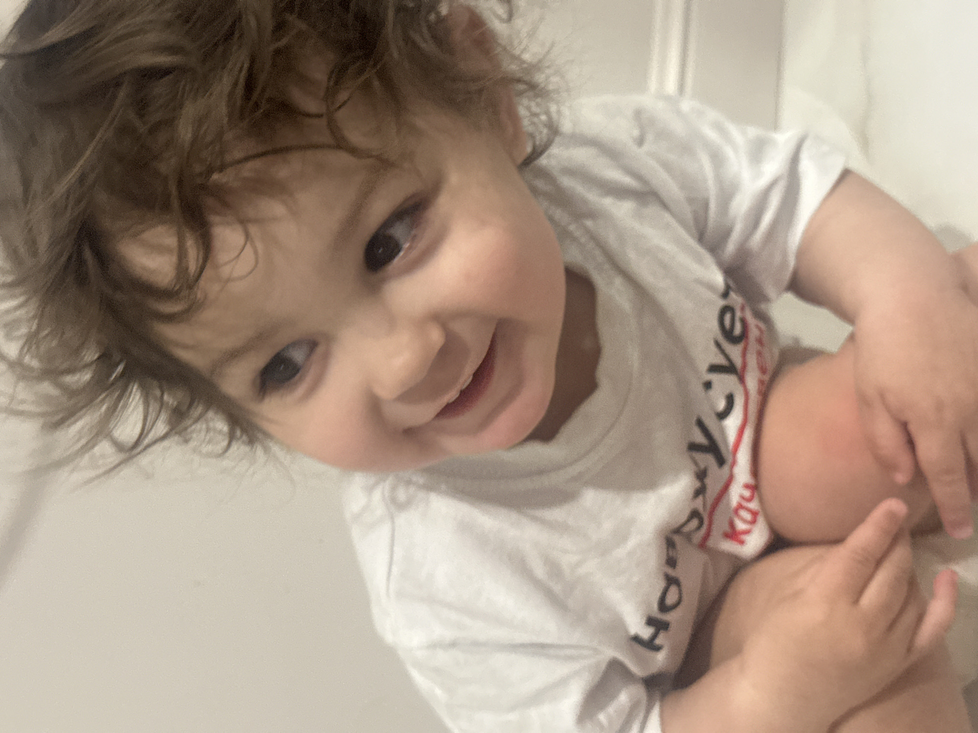 The image presents a close-up, high-angle portrait of a young child, likely a toddler, captured in an indoor setting. The child has expressive dark eyes and curly brown hair, which is a prominent feature around their face. A wide, joyful smile is directed towards the camera, revealing some teeth. The child's head is tilted, conveying a playful and engaging demeanor.
The child is wearing a white short-sleeved t-shirt. Visible text on the t-shirt includes "Ho" in black capital letters and "Ka" in red capital letters, positioned vertically on the chest. The child's body is partially visible, suggesting they are in a crouching or kneeling position with their hands resting on their knees.
The background is uniformly light-colored and out of focus, indicating an indoor environment without specific distinguishing features. The lighting is soft and even, highlighting the child's features. The overall impression is one of happiness and innocence.
Provided context indicates the location is Khimki, Russia.