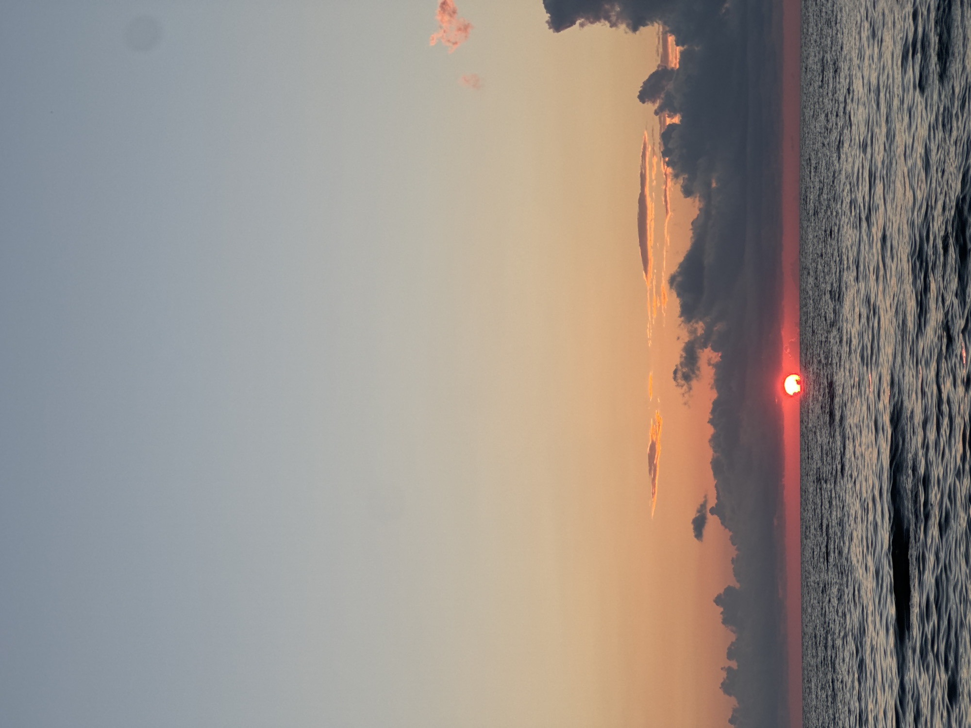 A vertical, full-frame shot depicts a sunset over the Black Sea in Batumi, Georgia. The sun, a bright red orb, is partially submerged below the horizon, casting a warm orange glow across the sky. Dark, dramatic clouds fill the middle ground, with lighter, wispy clouds scattered higher up. The water in the foreground is a deep blue-gray, with gentle ripples reflecting the muted colors of the sky. No people are visible in the frame.